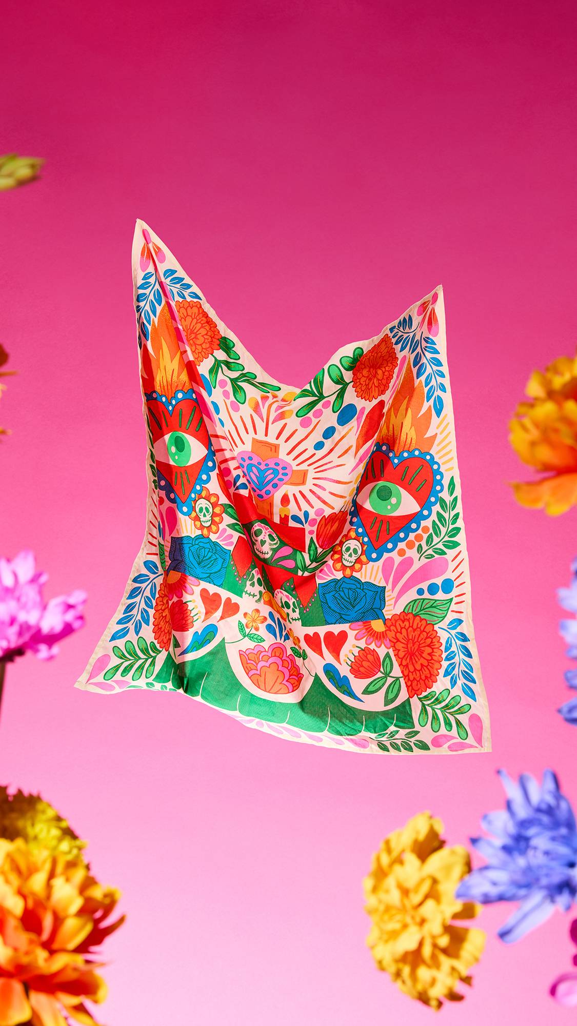 The image shows the vibrantly coloured, Día De Muertos-themed Amor Eterno fabric knot wrap. The wrap is suspended mid-air on a vibrant pink background with blurred marigold flowers around the edge. 