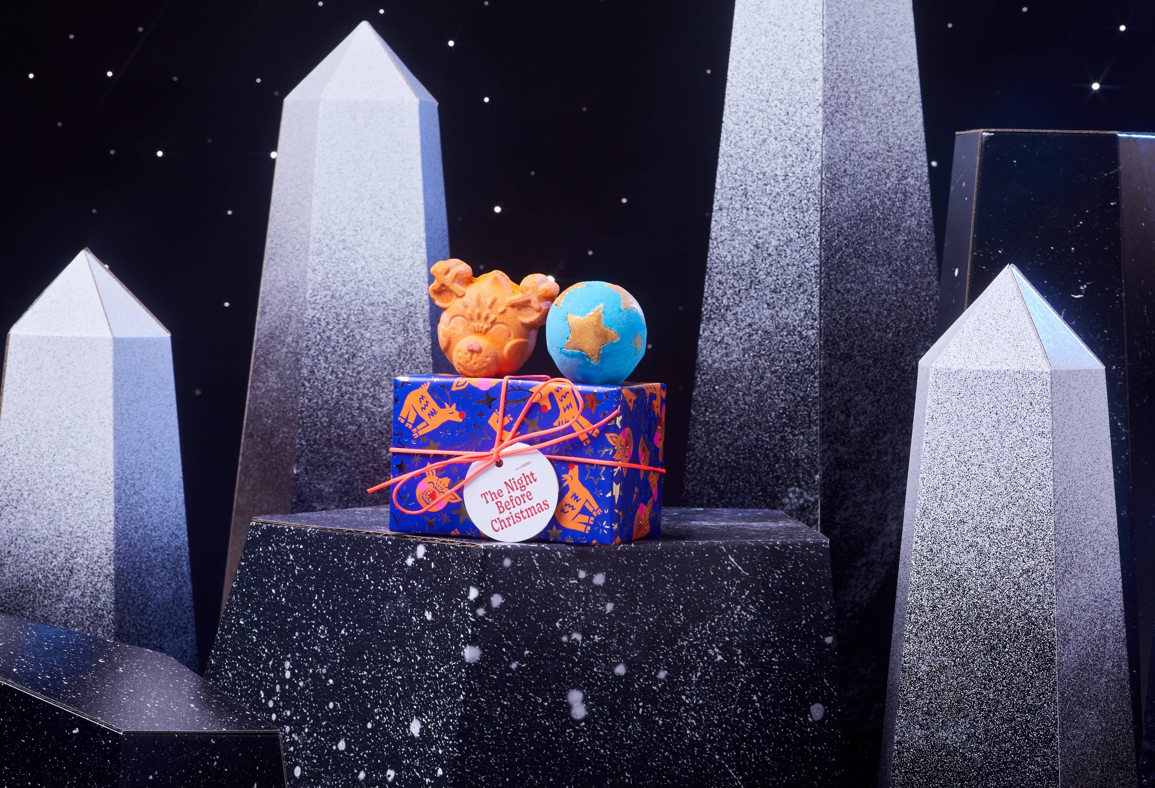 Night Before Christmas gift box and two bath bombs are on a dark chromatic platform among geometric shapes. 
