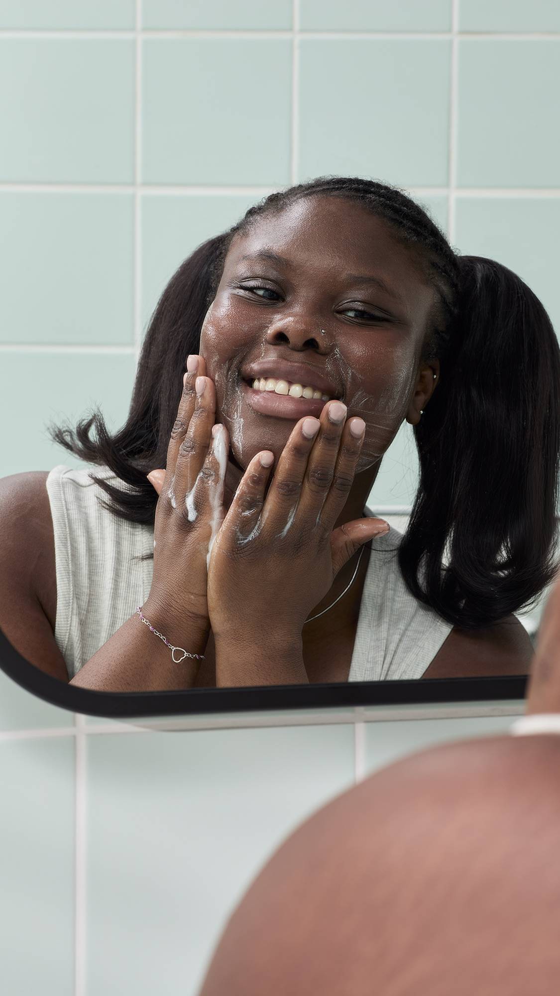A reflection in a bathroom mirror of a smiling model with dark hair in bunches, applying a white foaming lather to their face, created by the "Baby Bear" gummy facial cleanser, against pale green tiles.