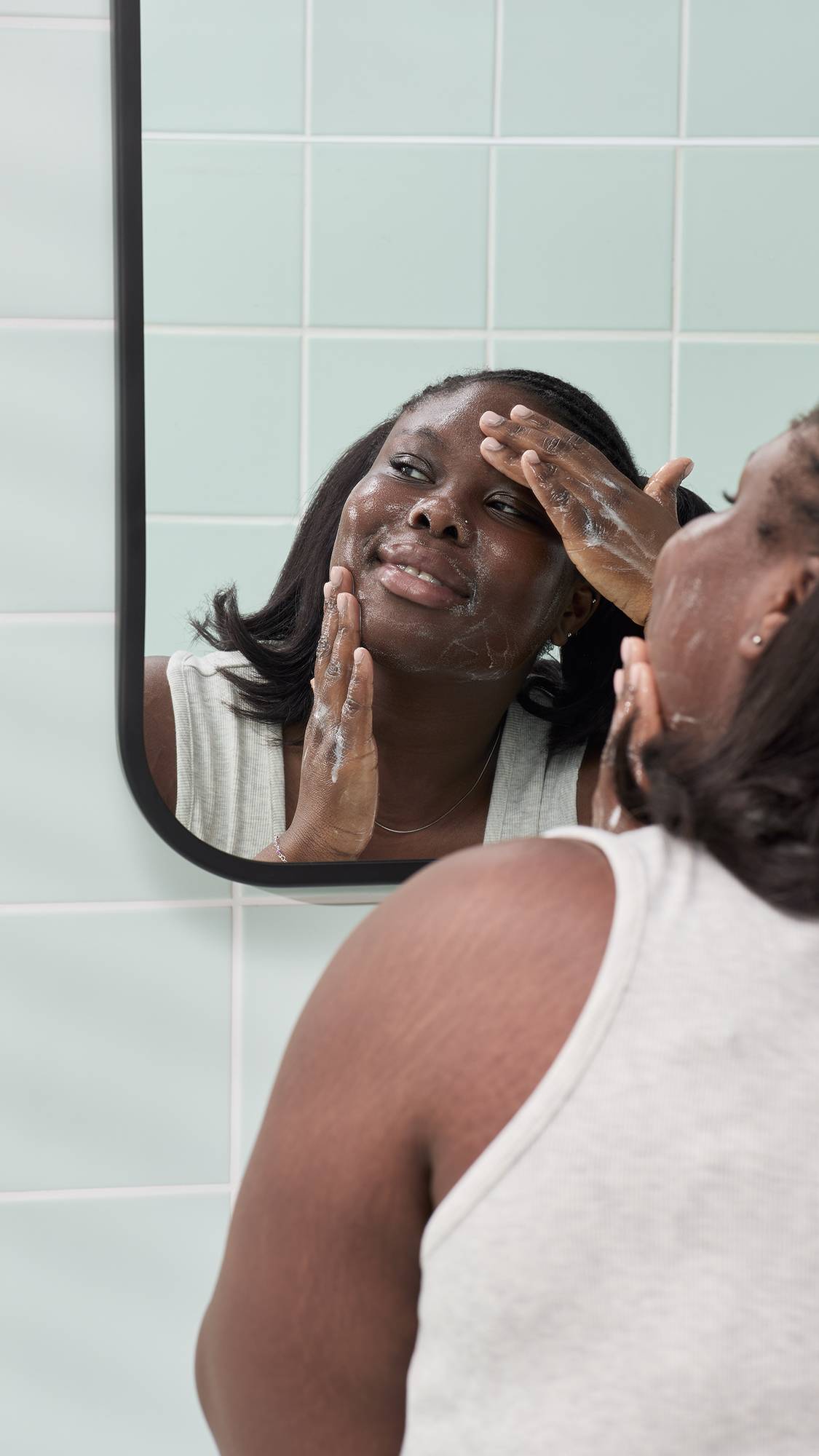A reflection in a bathroom mirror of a smiling model with dark hair in bunches, applying a white foaming lather to their face, created by the "Baby Bear" gummy facial cleanser, against pale green tiles.