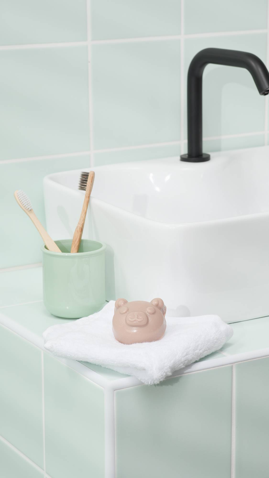 A pale, bear-shaped gummy facial cleanser rests on a folded white flannel by a white square basin. A light green cup holding two bamboo toothbrushes and a black tap are visible against pale green wall tiles.