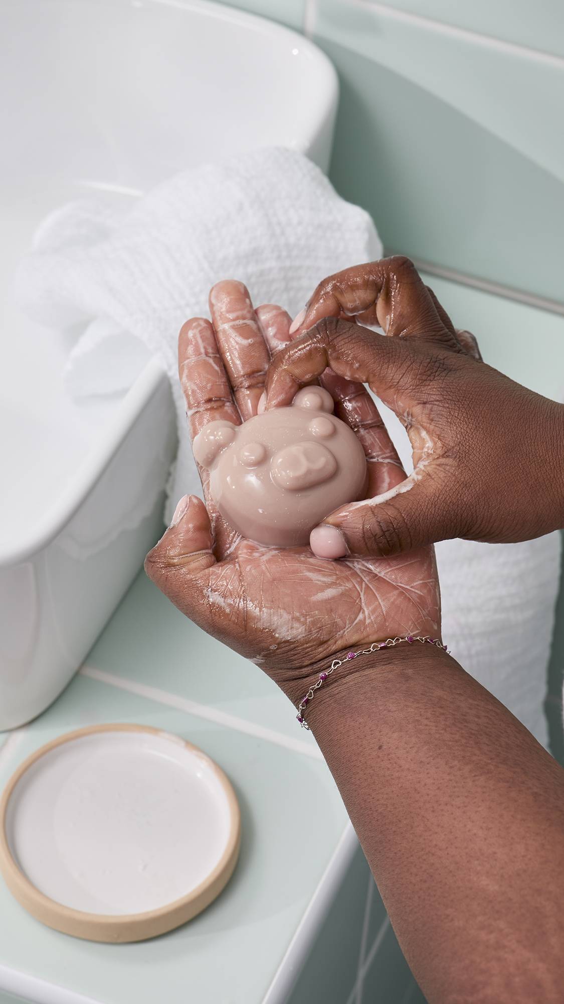 A model holds a small, bear-shaped facial jelly in their hands to create a lather. The "Baby Bear" facial jelly has a simple, rounded face. In the background, a white sink, a towel, and a ceramic soap dish are visible against pale green wall tiles.