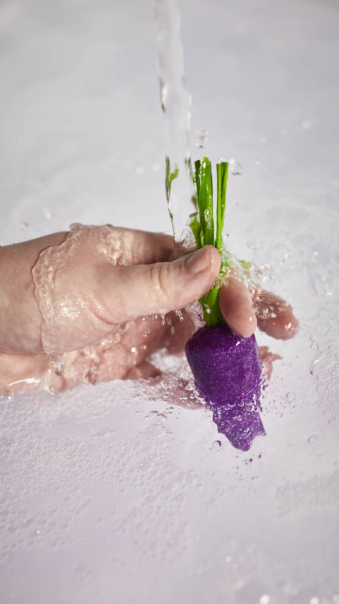 A close-up image of the model's hand holding the purple carrot bubble bar by the green leaves under running tap water into the bath below. 
