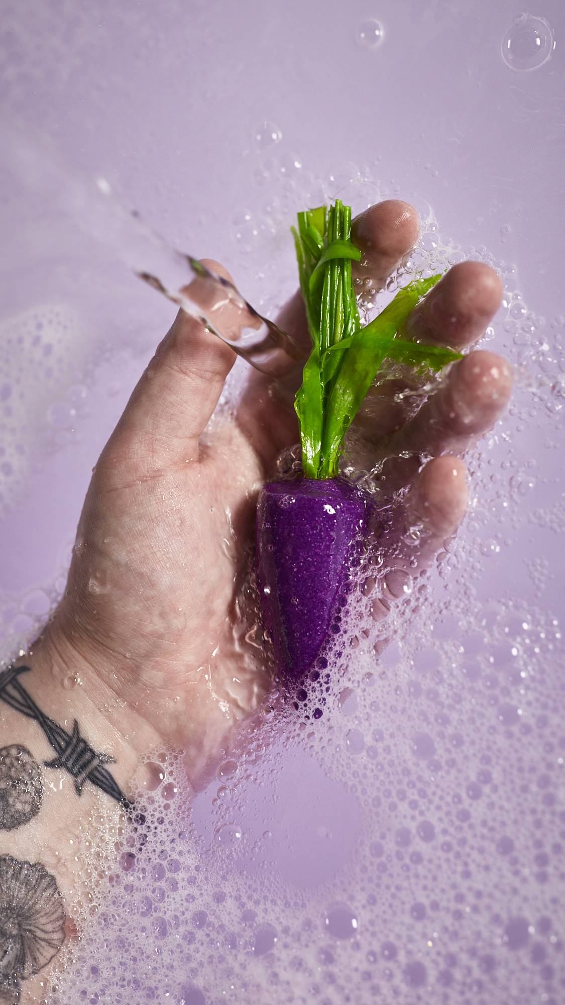 A close-up image of the model's hand holding the purple carrot bubble bar under running water creating delicate, bubbly water below. 