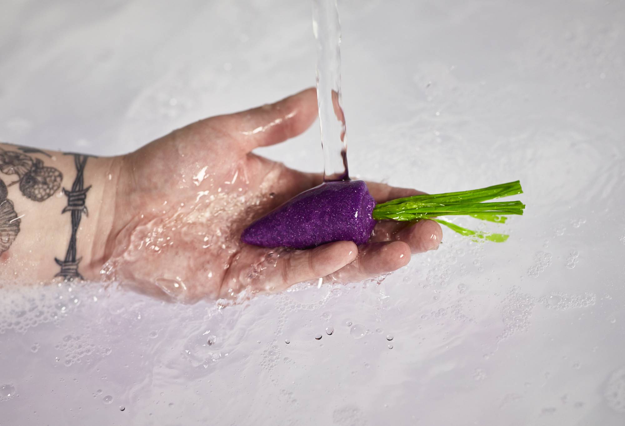 A close-up image of the model's hand holding the purple carrot bubble bar under running water.