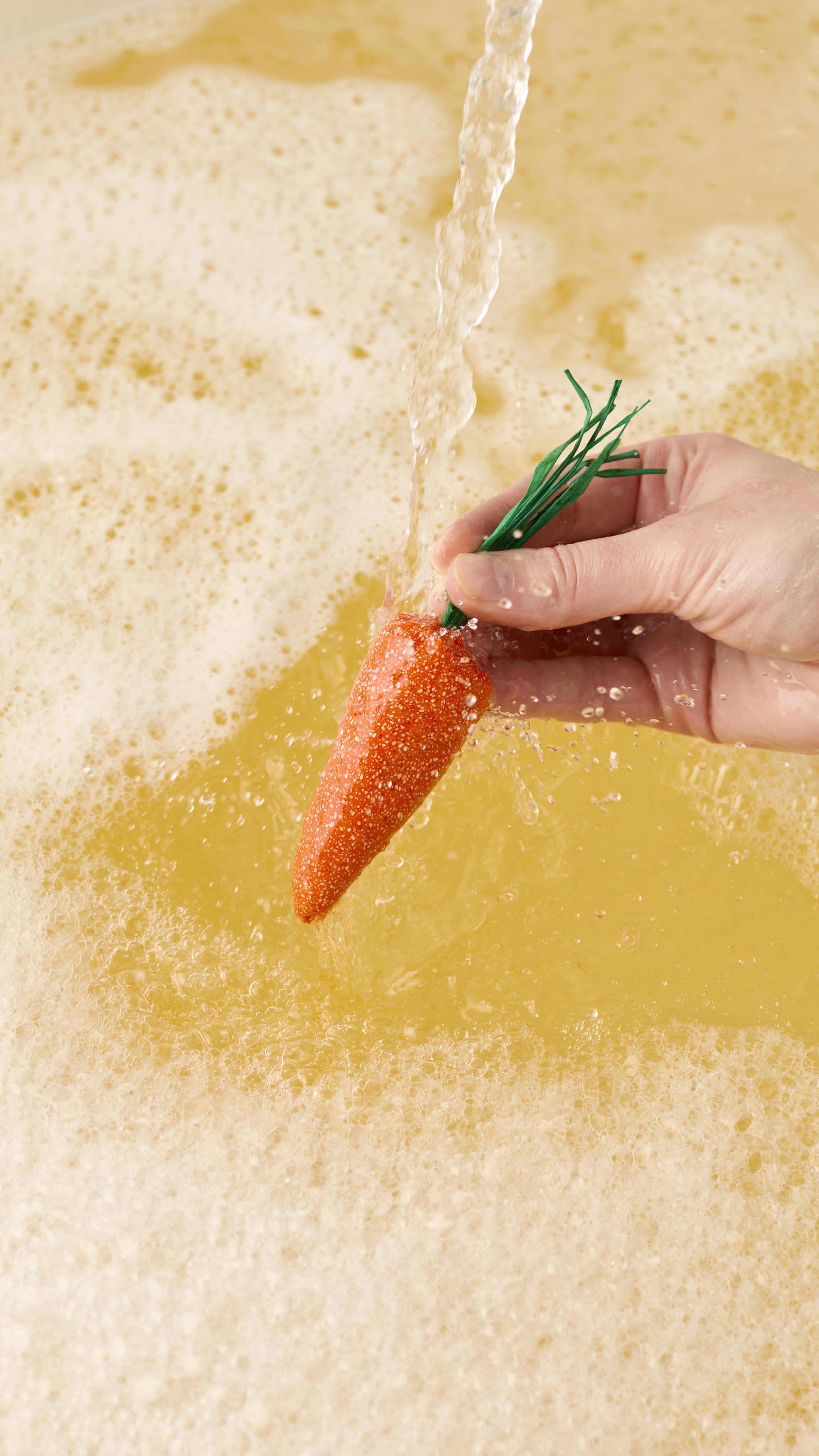 A hand holds a bright orange, carrot-shaped bubble bar with green raffia stems under a stream of running water, creating pale yellow bathwater and a thick layer of frothy white bubbles.