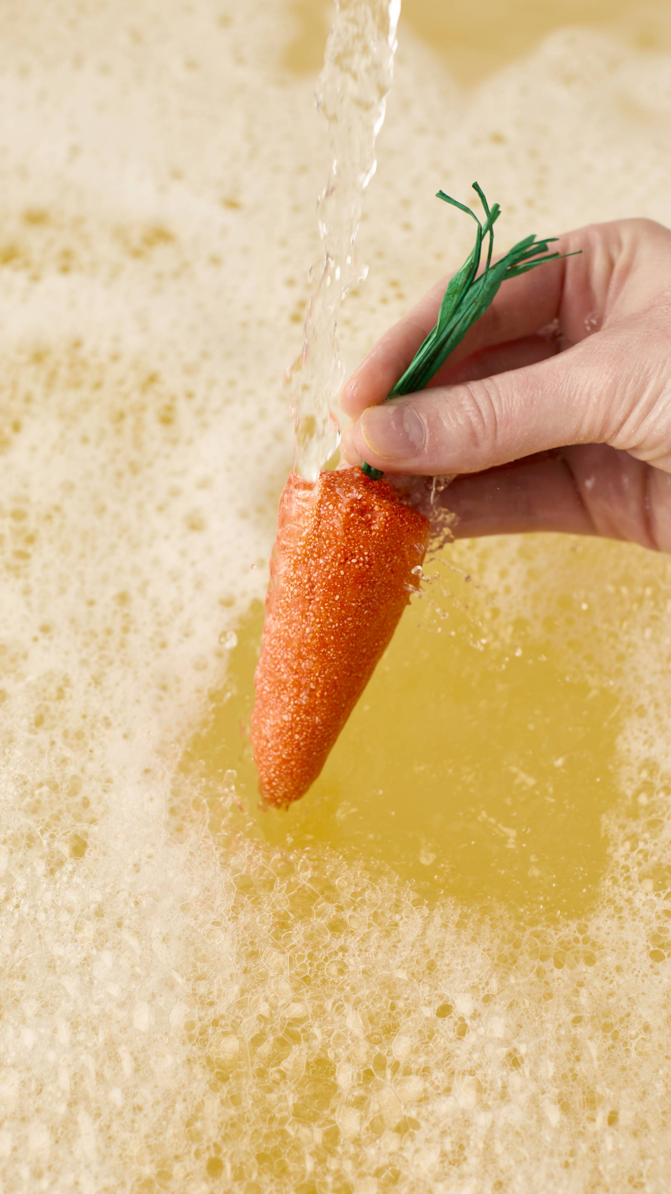 The image shows a close-up of a model's hand holding an orange "Baby Rainbow Carrot" bubble bar by its green paper stalks. The bubble bar is held under a stream of running water over a bathtub filled with thick white bubbles and golden water.