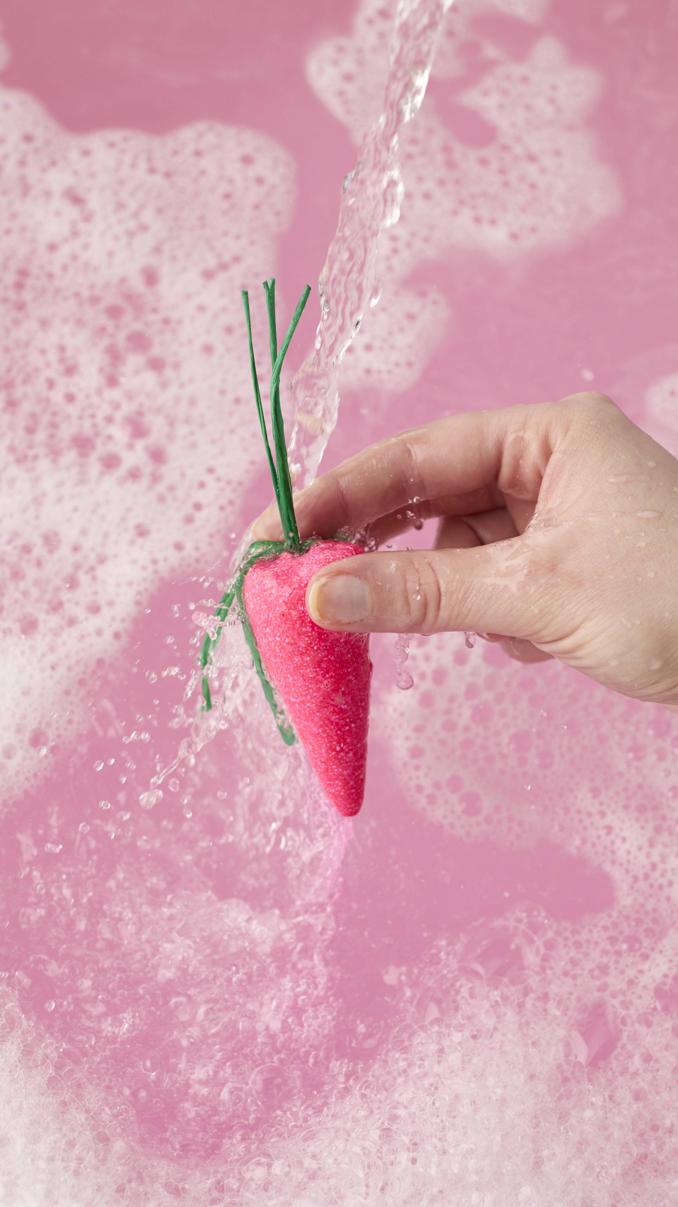 A hand holds a bright pink, carrot-shaped bubble bar with green raffia stems under a stream of running water. The bar creates a pool of vibrant pink bathwater topped with a thick layer of frothy white bubbles.