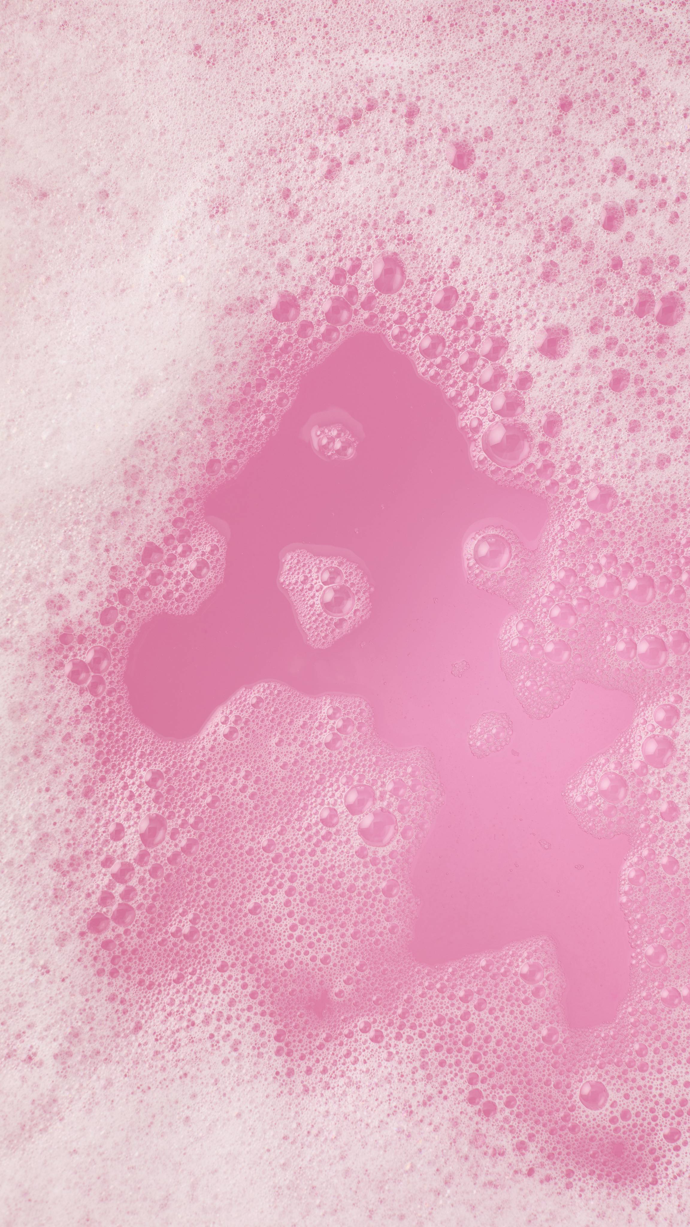 A top-down view of vibrant pink bathwater with an island of thick, frothy white bubbles. Small clusters of foam drift across the clear pink surface, creating a soft, indulgent texture.