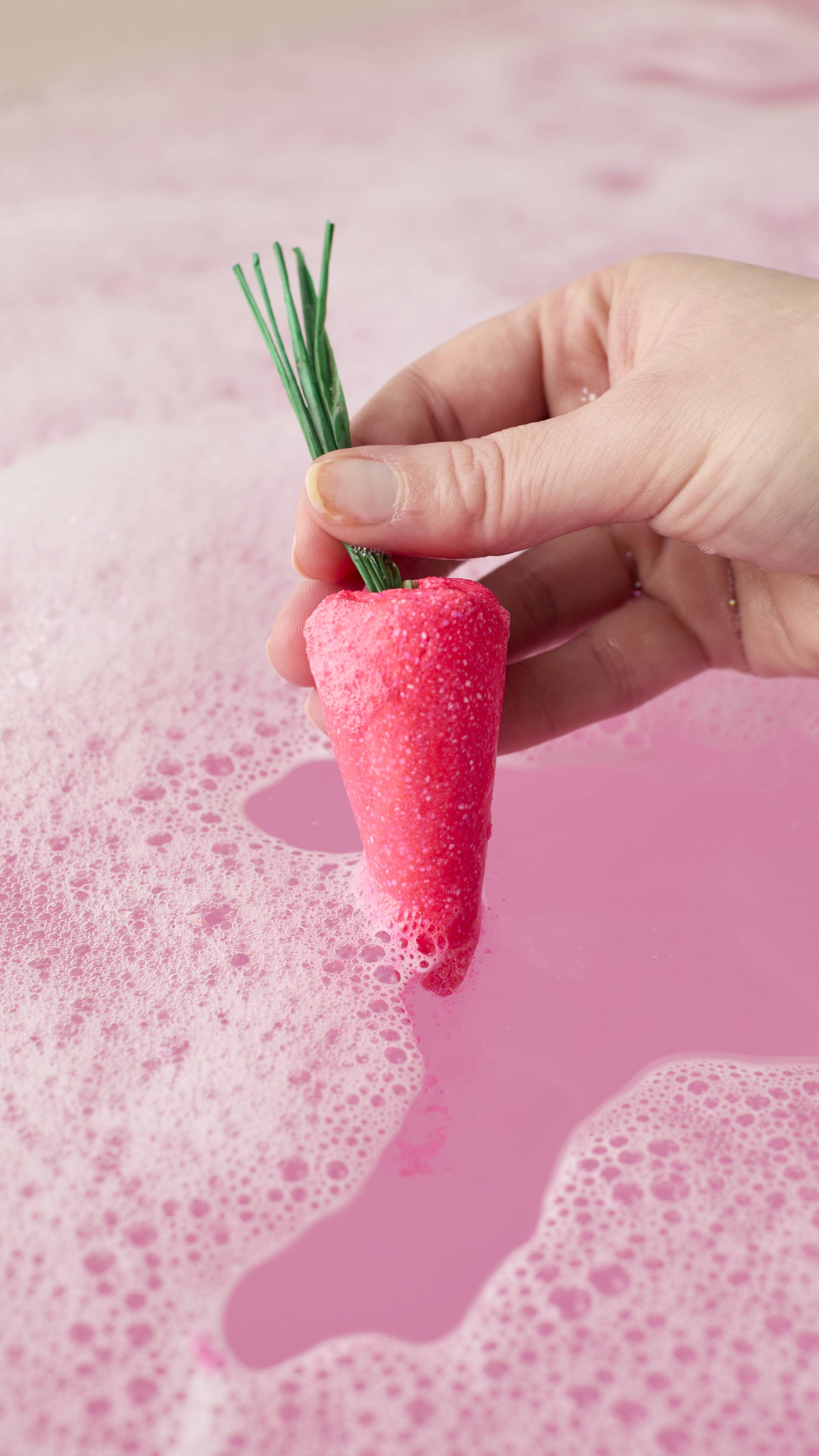 A hand holds a bright pink, carrot-shaped bubble bar with green stems over a pool of vibrant pink bathwater. Thick white bubbles are beginning to form on the surface around the carrot as it dips into the water.