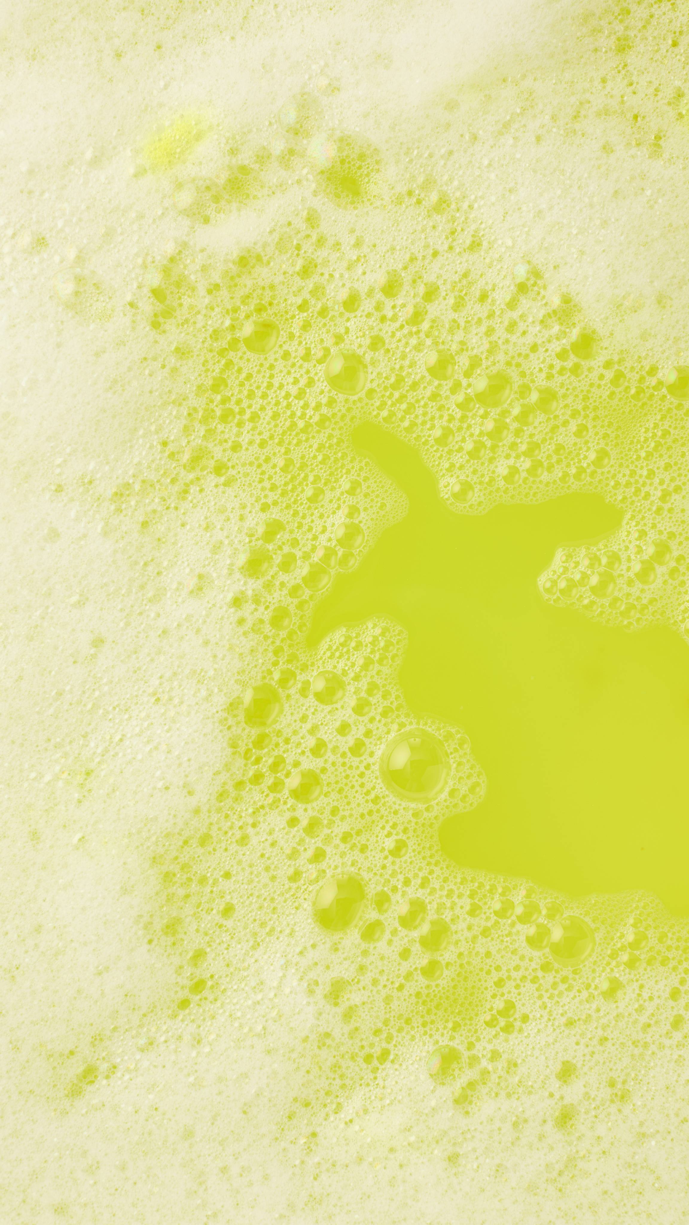 A top-down view of vibrant yellow bathwater with an island of thick, frothy white bubbles. Small clusters of foam drift across the clear yellow surface, creating a soft, indulgent texture.