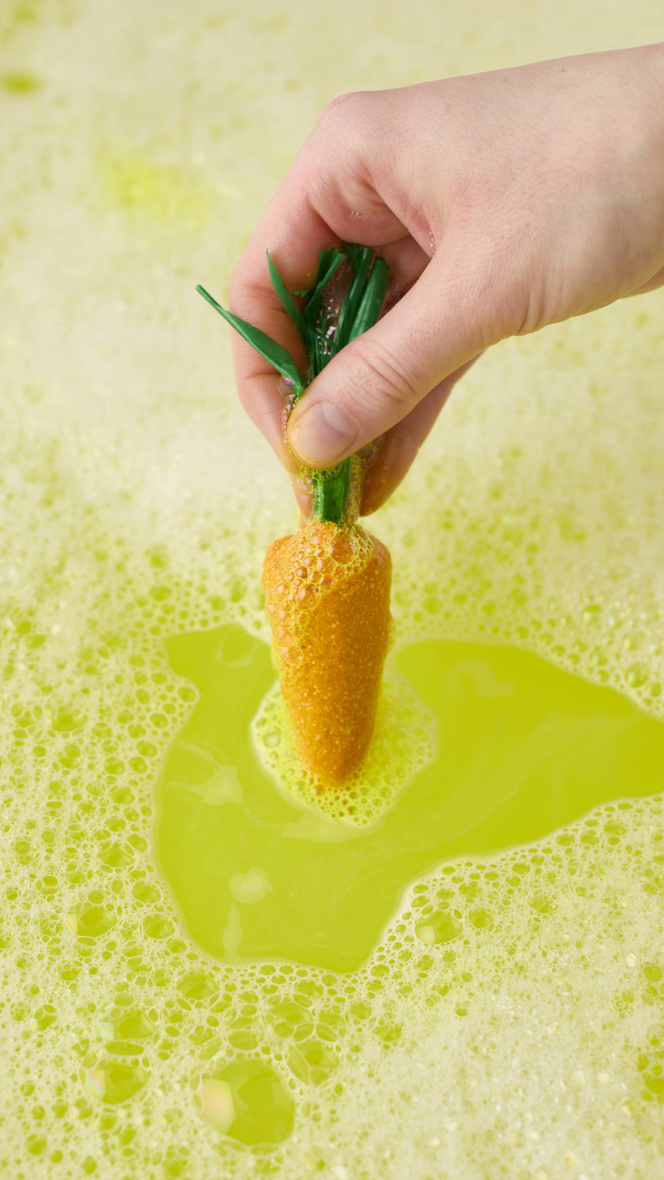A hand holds a bright yellow carrot-shaped bubble bar by its green stems, dipping it into lime-green bathwater. Thick white foam and yellow bubbles gather around the carrot's base as it creates a vibrant bath.