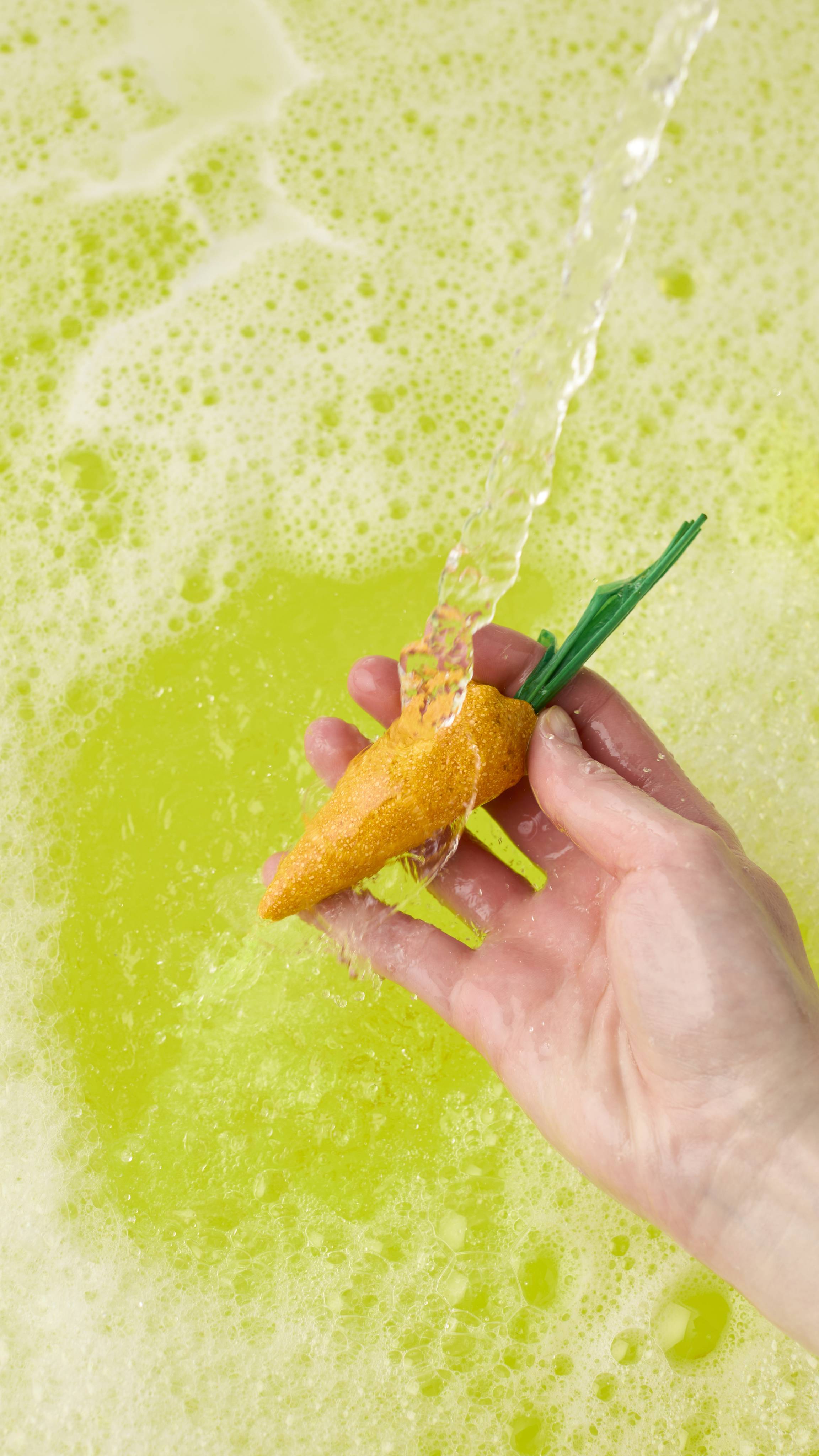 A hand holds a bright yellow, carrot-shaped bubble bar with green raffia stems under a stream of running water. The bar dissolves into vibrant, lime-green bathwater topped with a thick layer of frothy white bubbles.