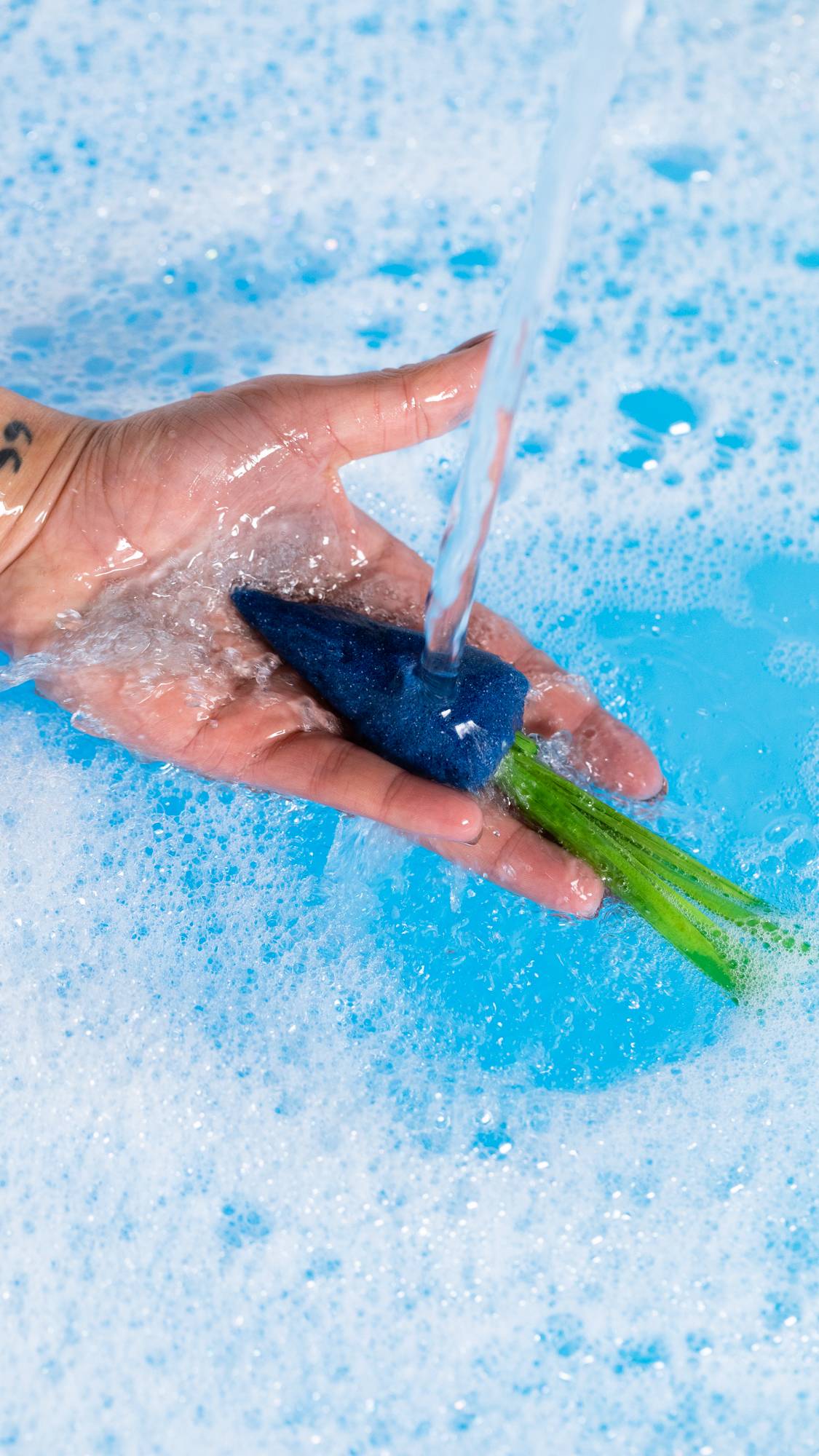 A close-up image of the model's hand holding the blue carrot bubble bar under running water creating a deep, bubble, blue sea below. 