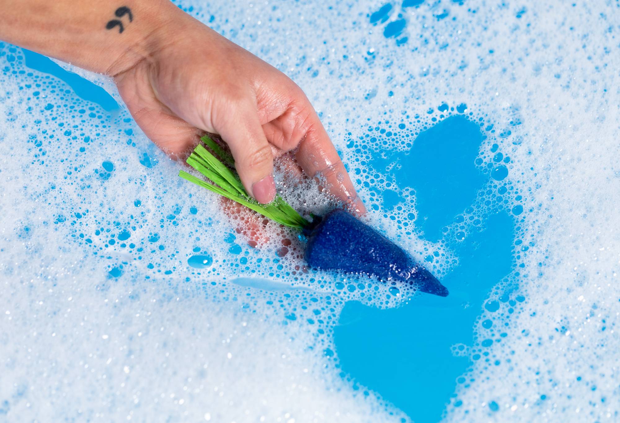 A close-up of the model holding the blue carrot bubble bar in the blue bath water surrounded by thick bubbles. 