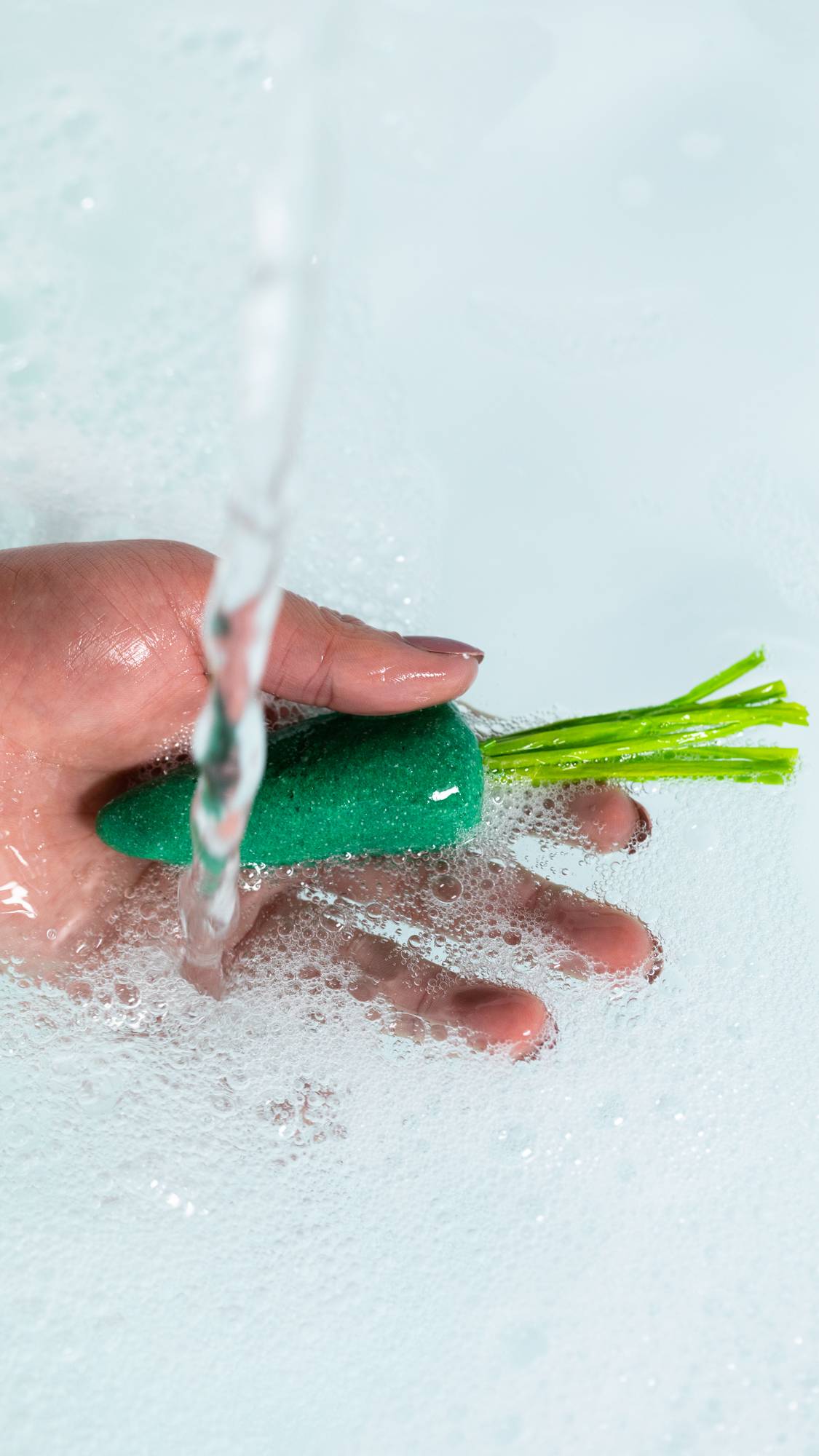 A close-up image of the model's hand holding the green carrot bubble bar under running water creating fresh, bubbly water below. 