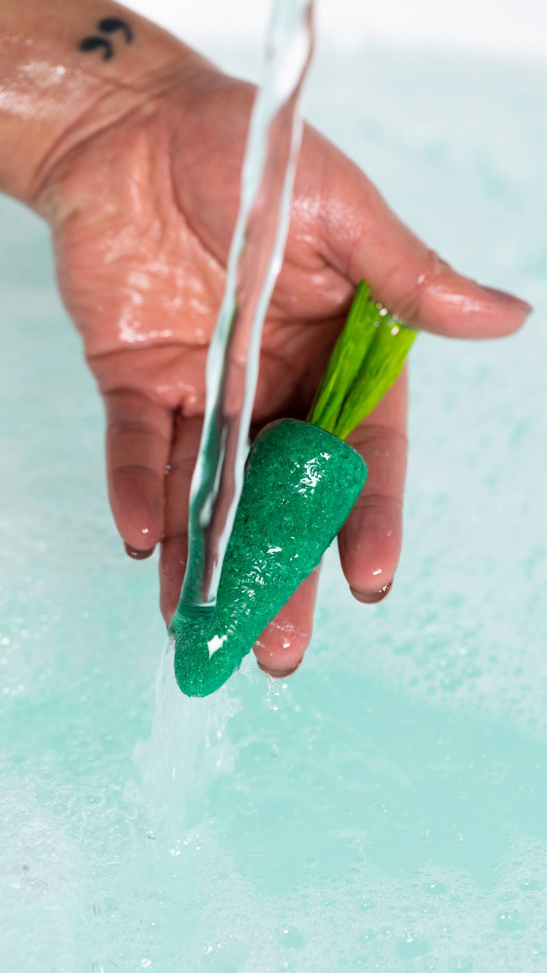 A close-up image of the model's hand holding the green carrot bubble bar under running water creating fresh, bubbly water below. 