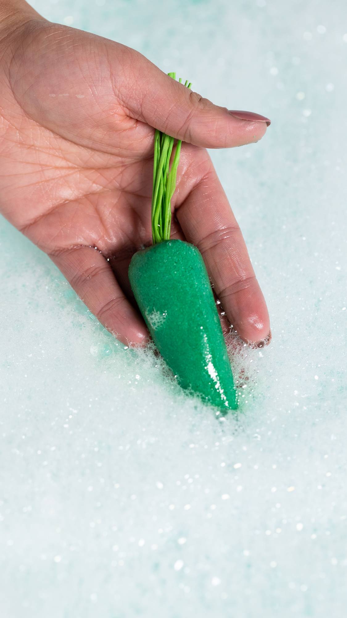 A close-up of the model holding the green carrot bubble bar in the blue bath water surrounded by thick bubbles. 