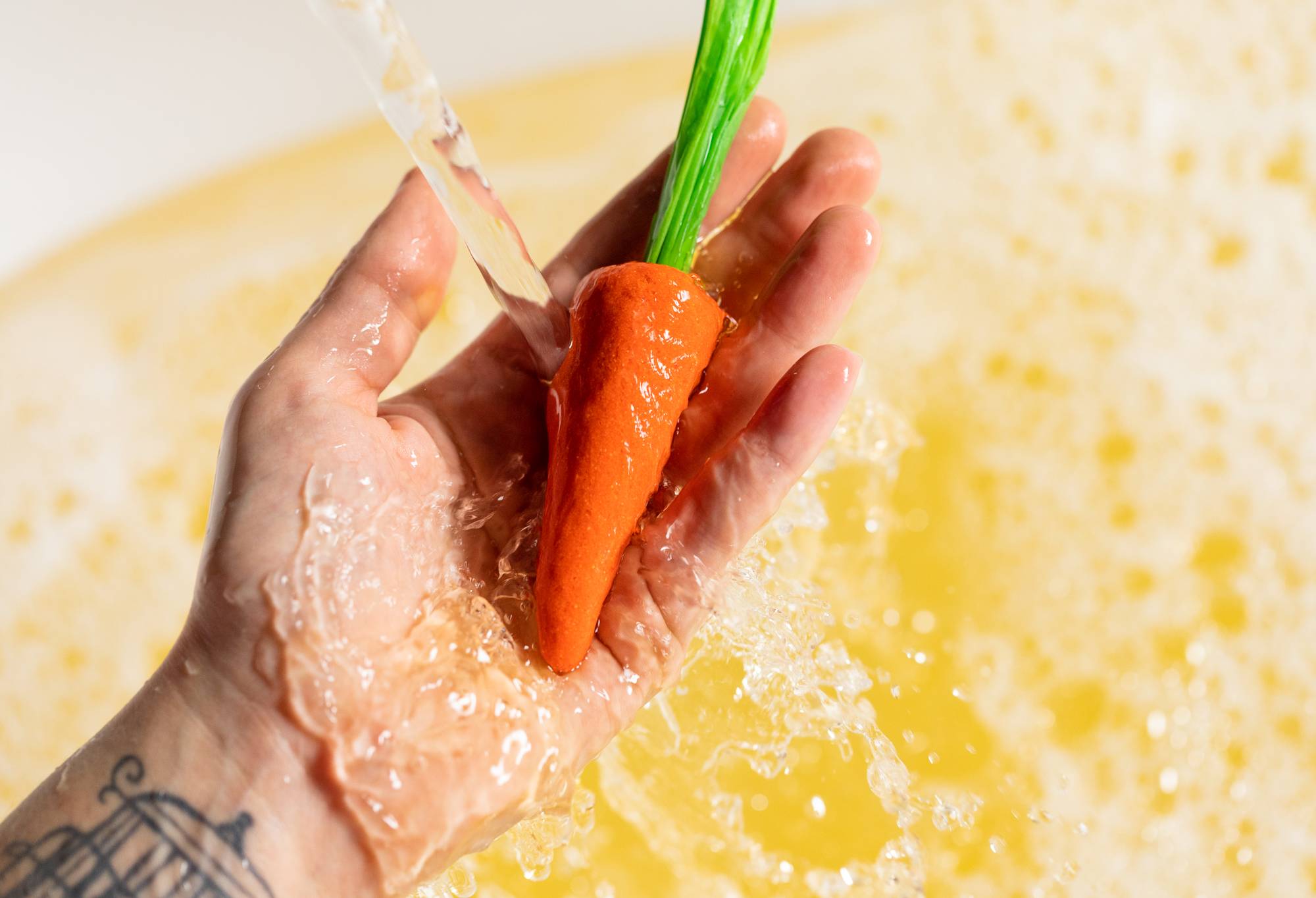 A close-up image of the model's hand holding the orange carrot bubble bar under running water creating light, bubbly water below. 