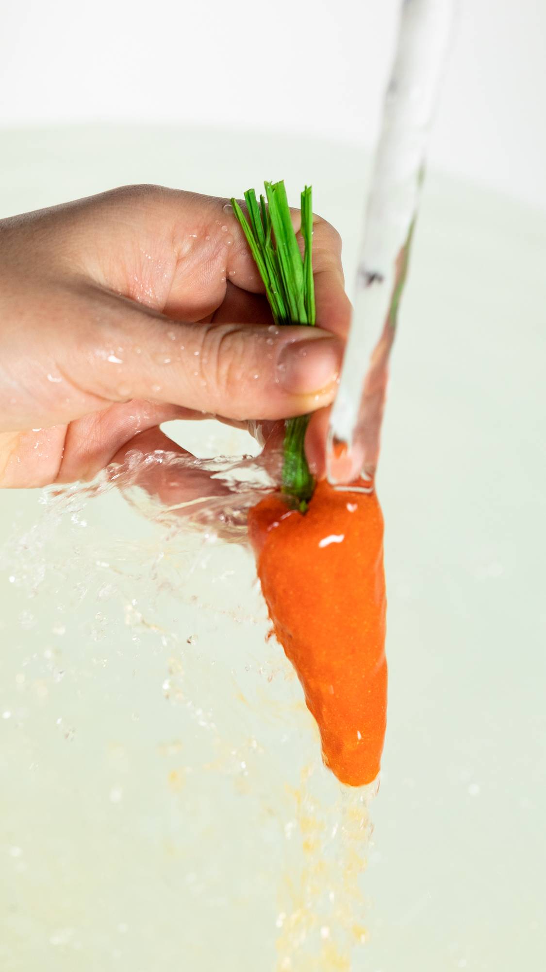 A close-up image of the model's hand holding the orange carrot bubble bar by the green leaves under running tap water into the bath below. 