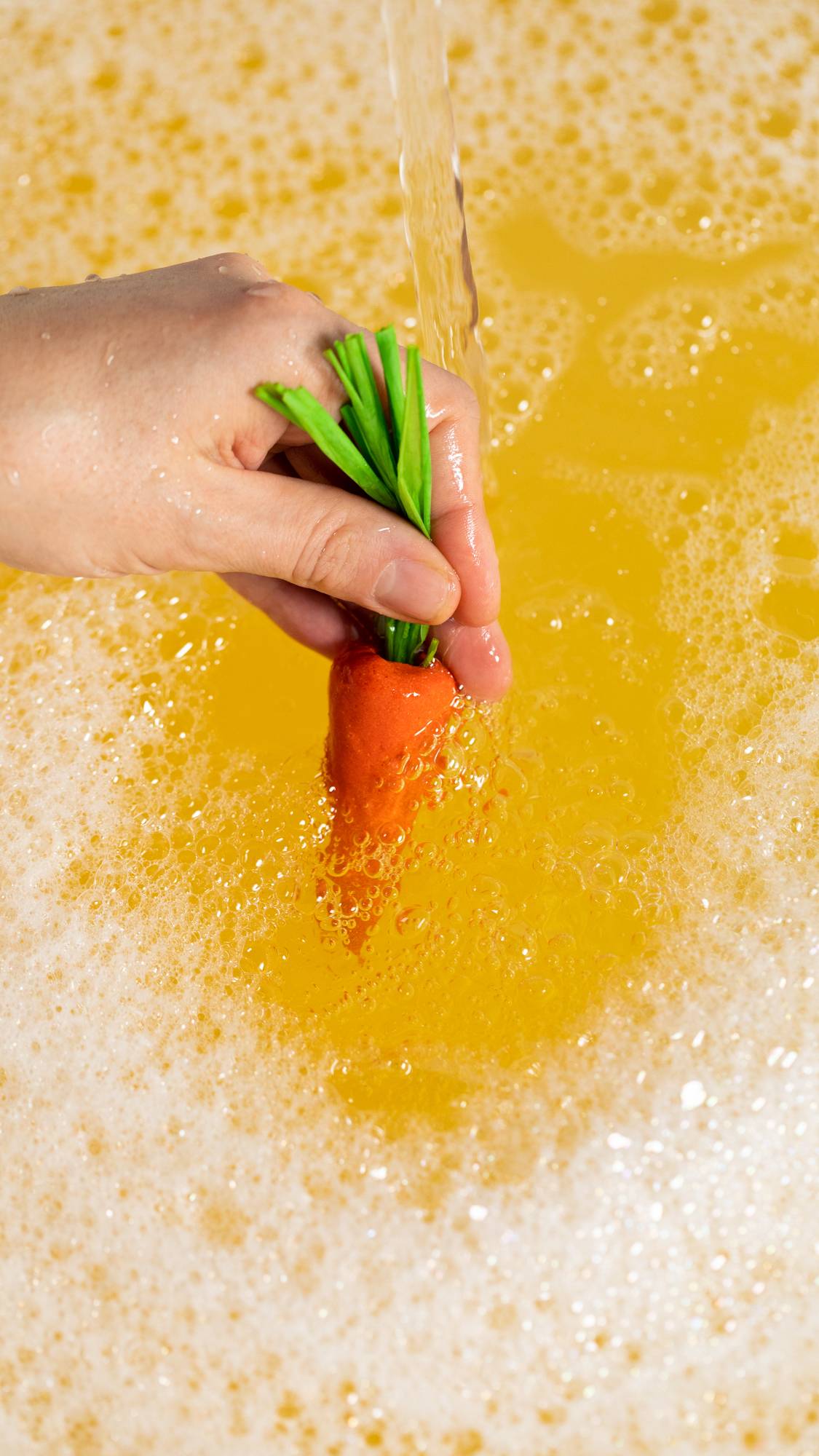 A close-up image of the model's hand dipping the orange carrot bubble bar into the bath under running water creating light, orange bubbles.