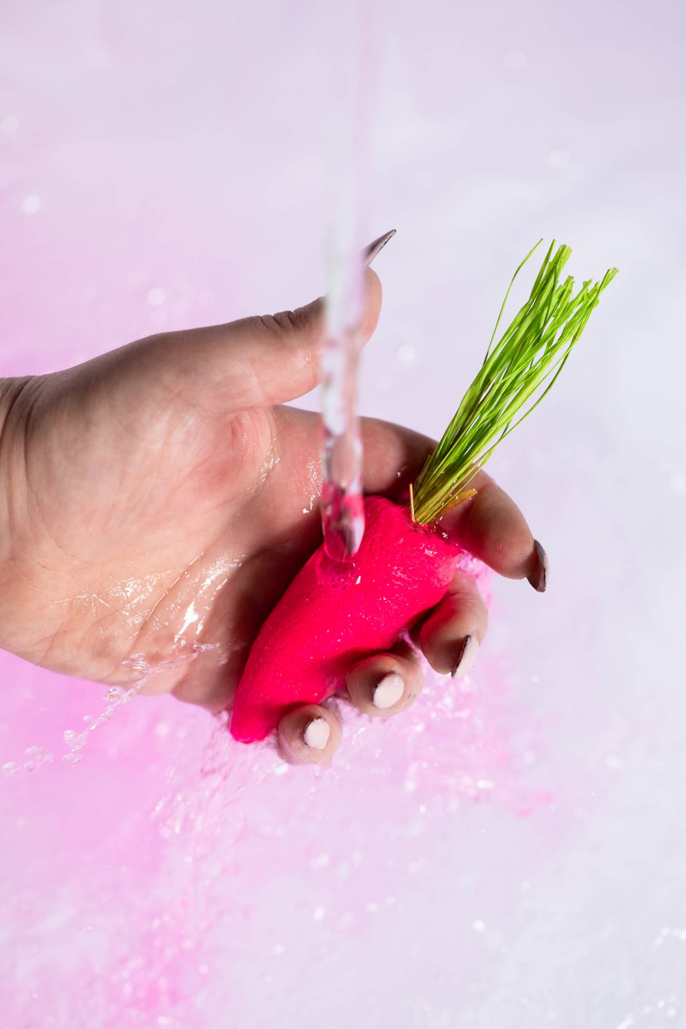 A close-up image of the model's hand holding the pink carrot bubble bar under running water creating light, bubbly water below. 