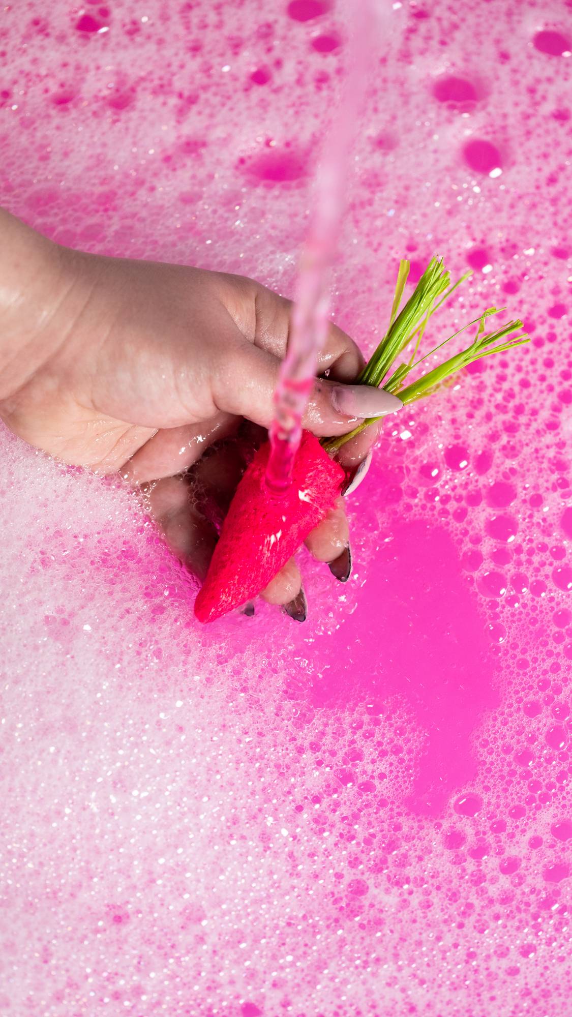 A close-up image of the model's hand holding the pink carrot bubble bar under running water creates a deep pink, bubbly bath. 