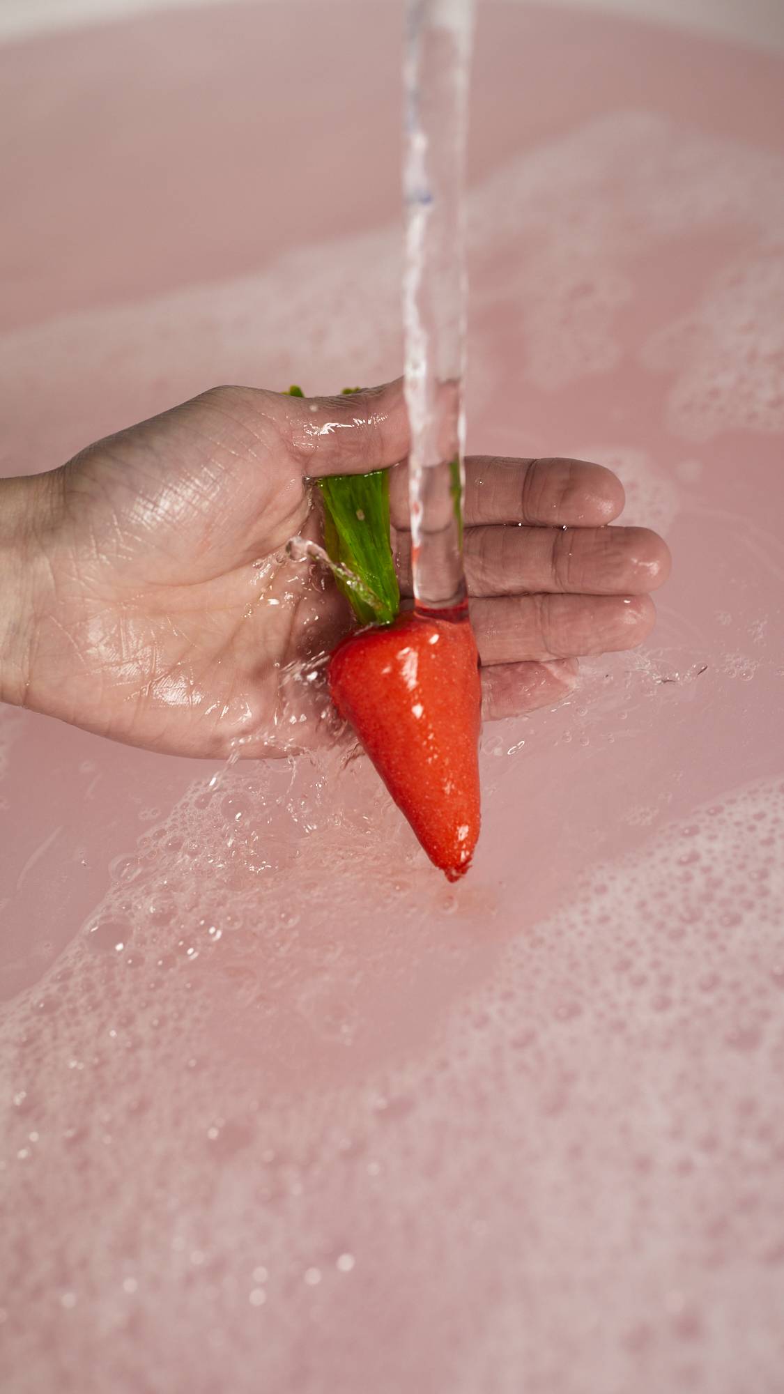 A close-up image of the model's hand holding the red carrot bubble bar by the green leaves under running tap water into the bath below. 
