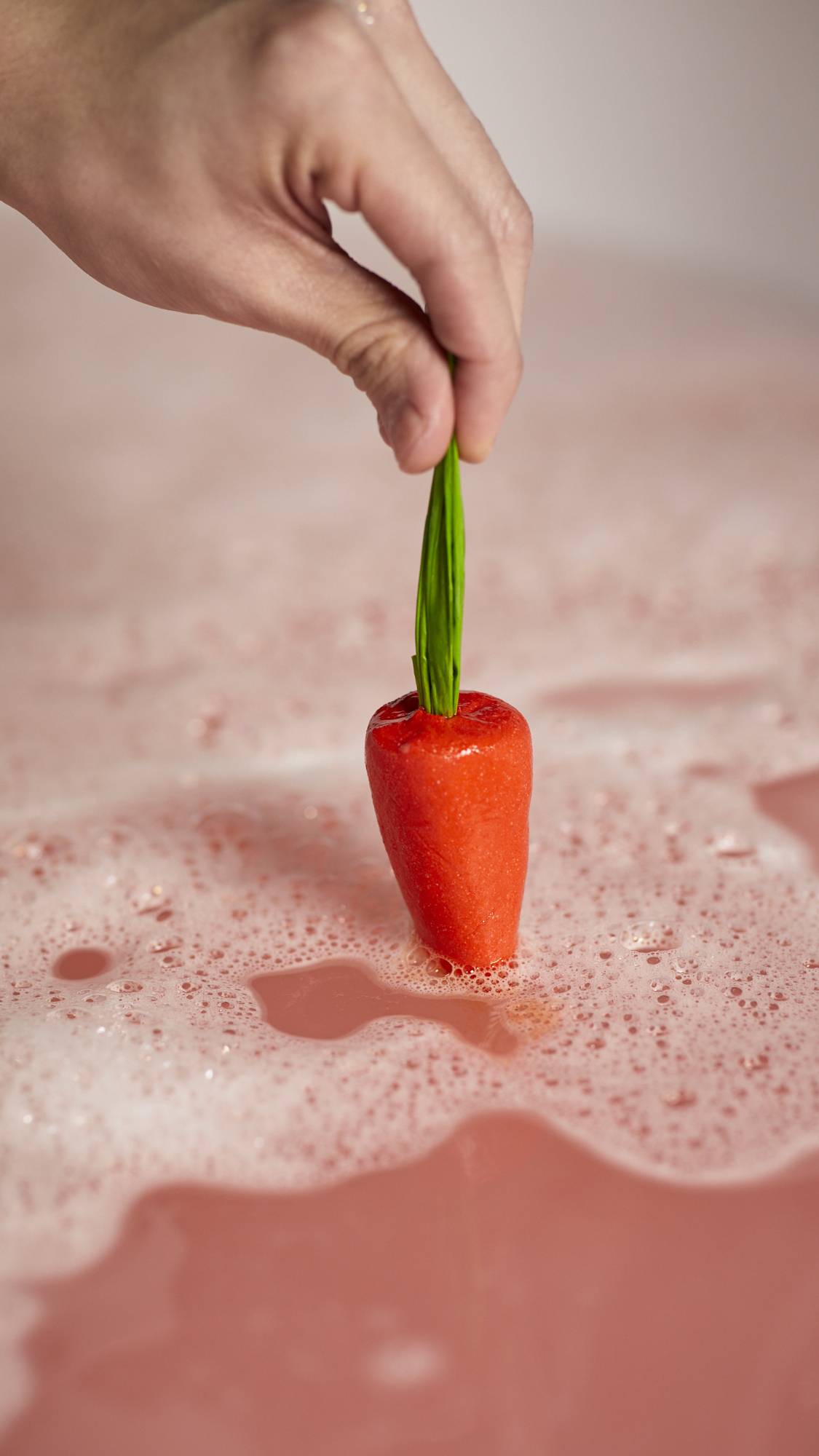 A close-up of the model holding the red carrot bubble bar by the tip of the green leaves dipping it into the bath water surrounded by thick bubbles. 