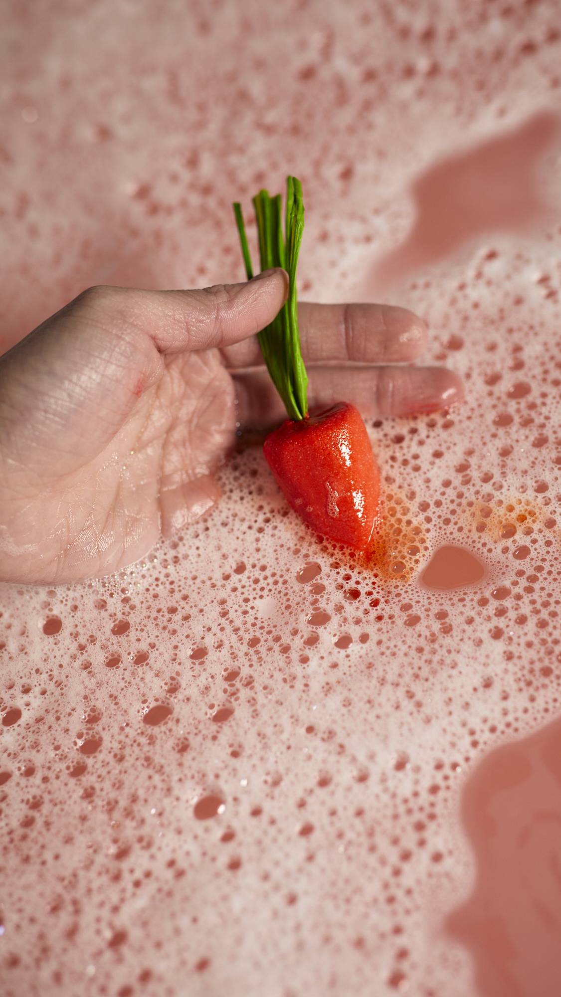 A close-up of the model holding the red carrot bubble bar in the blue bath water surrounded by thick bubbles. 