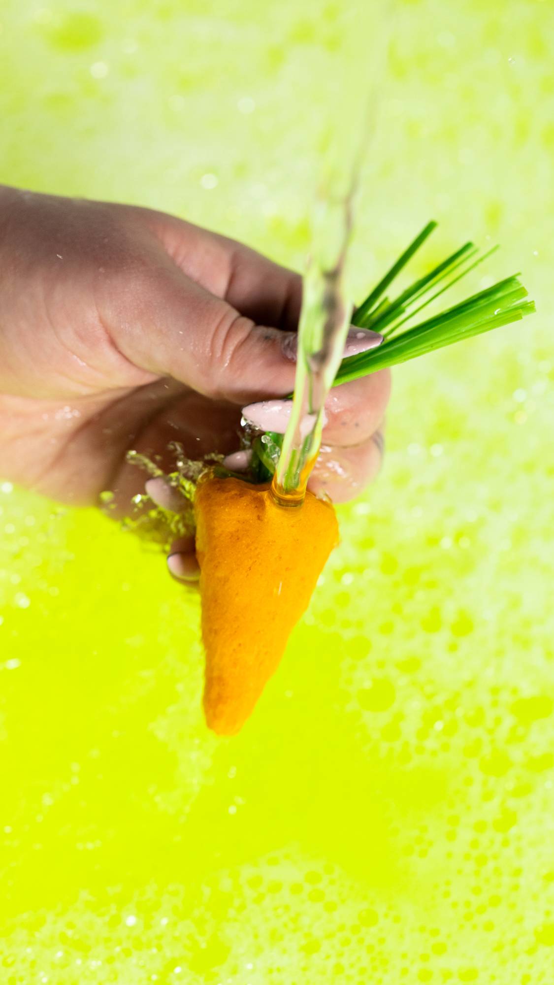 A close-up image of the model's hand holding the yellow carrot bubble bar by the green leaves under running tap water making a neon yellow bubble bath. below. 