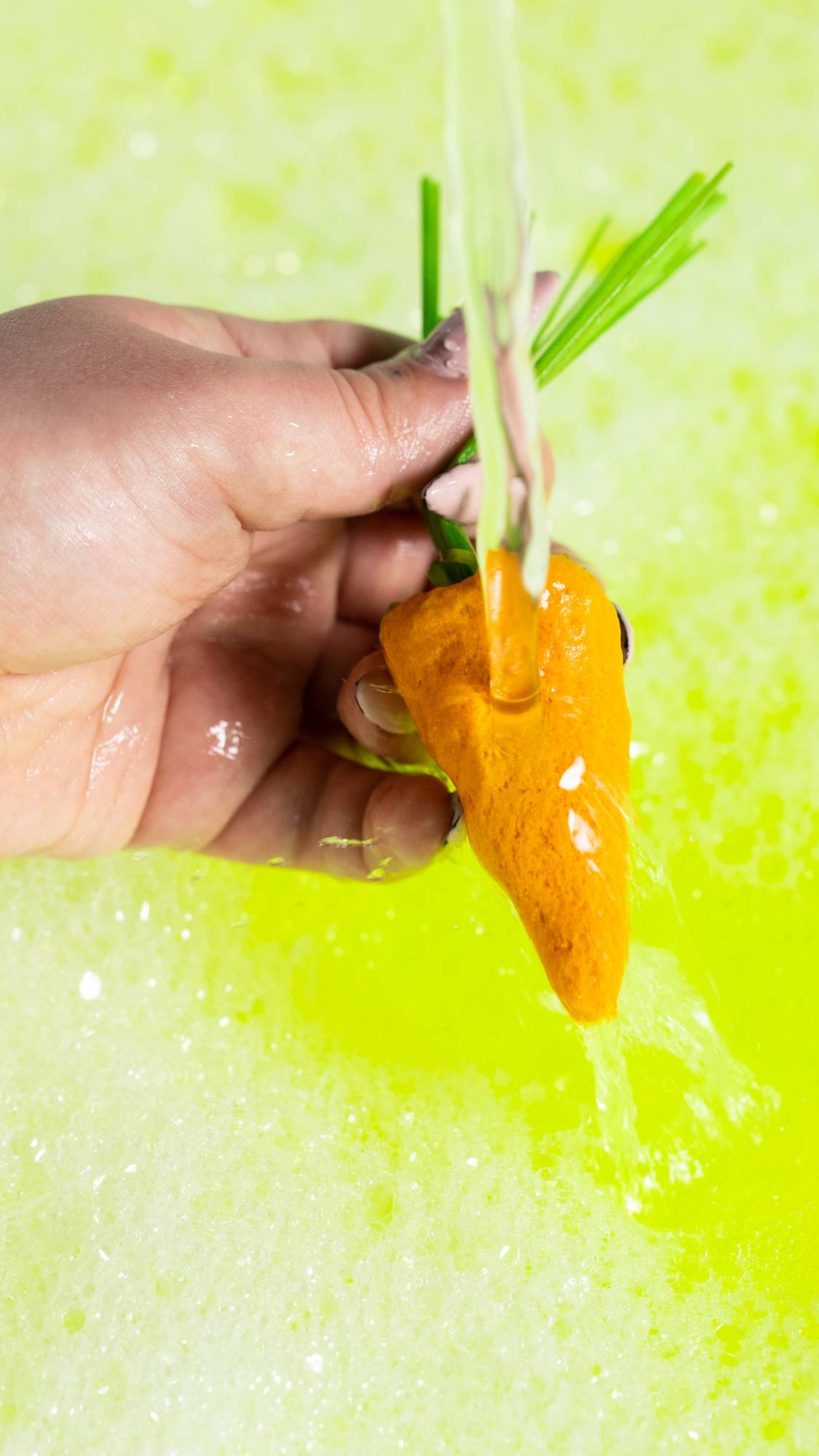 A close-up image of the model's hand holding the yellow carrot bubble bar by the green leaves under running tap water making a neon yellow bubble bath. below. 