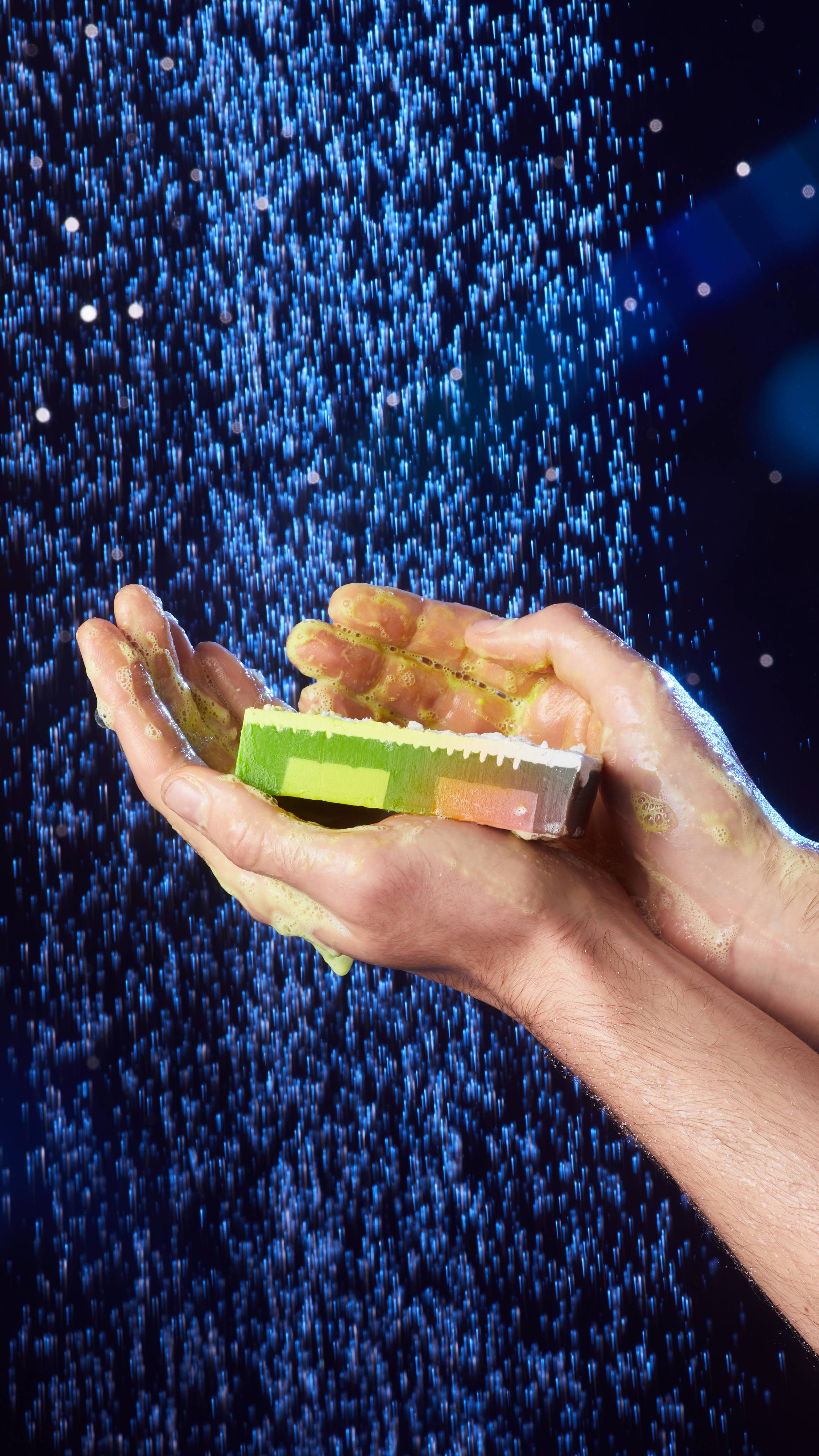 A close-up of the model's hands under shower water holding the Baked Alaska soap with thick, green lather.