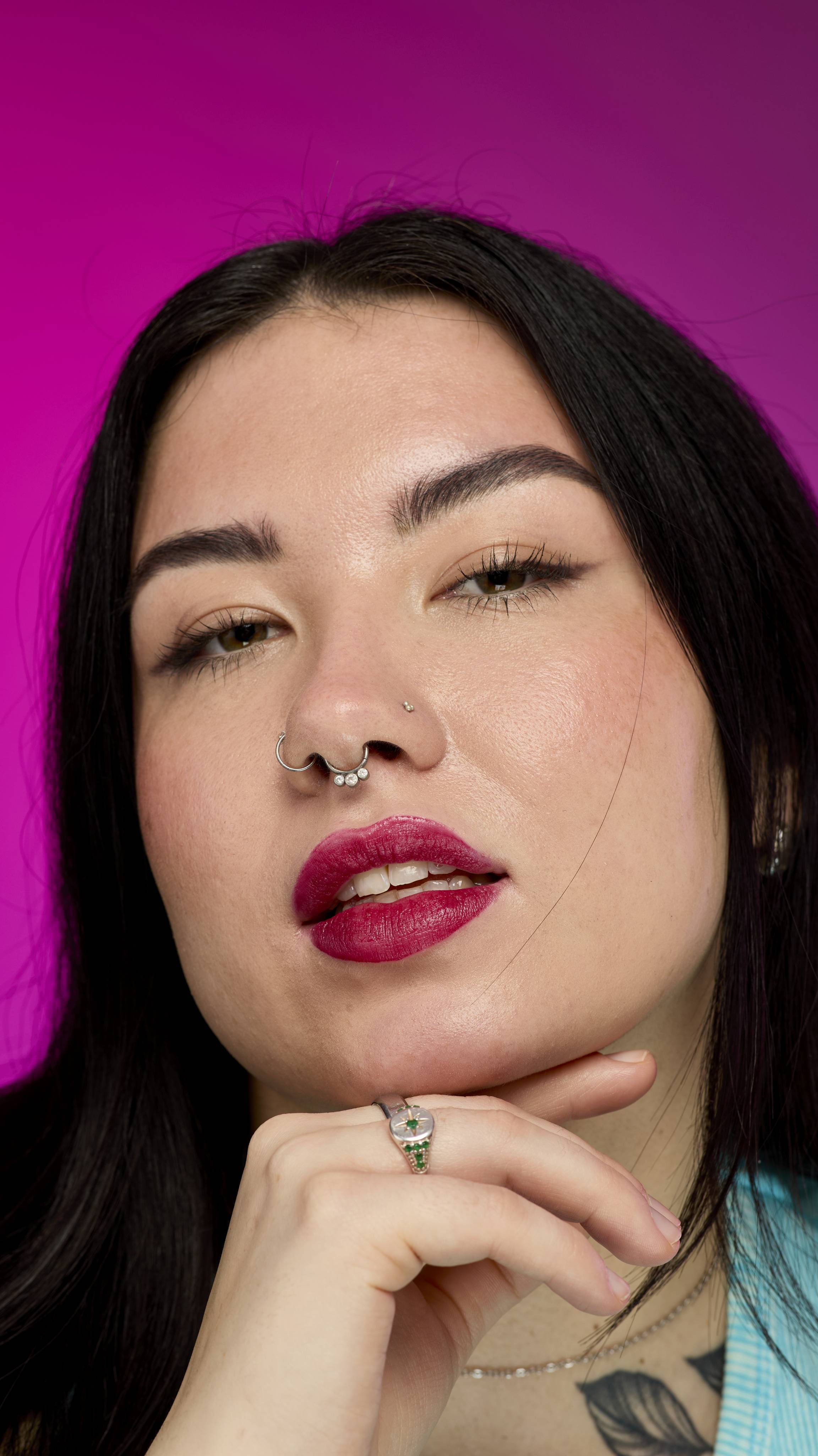 A smiling person with long, dark hair, wearing a yellow top, is shown from the chest up. They are on a vibrant pink background, showing off the deep red Banger lipstick with a subtle shimmer. 