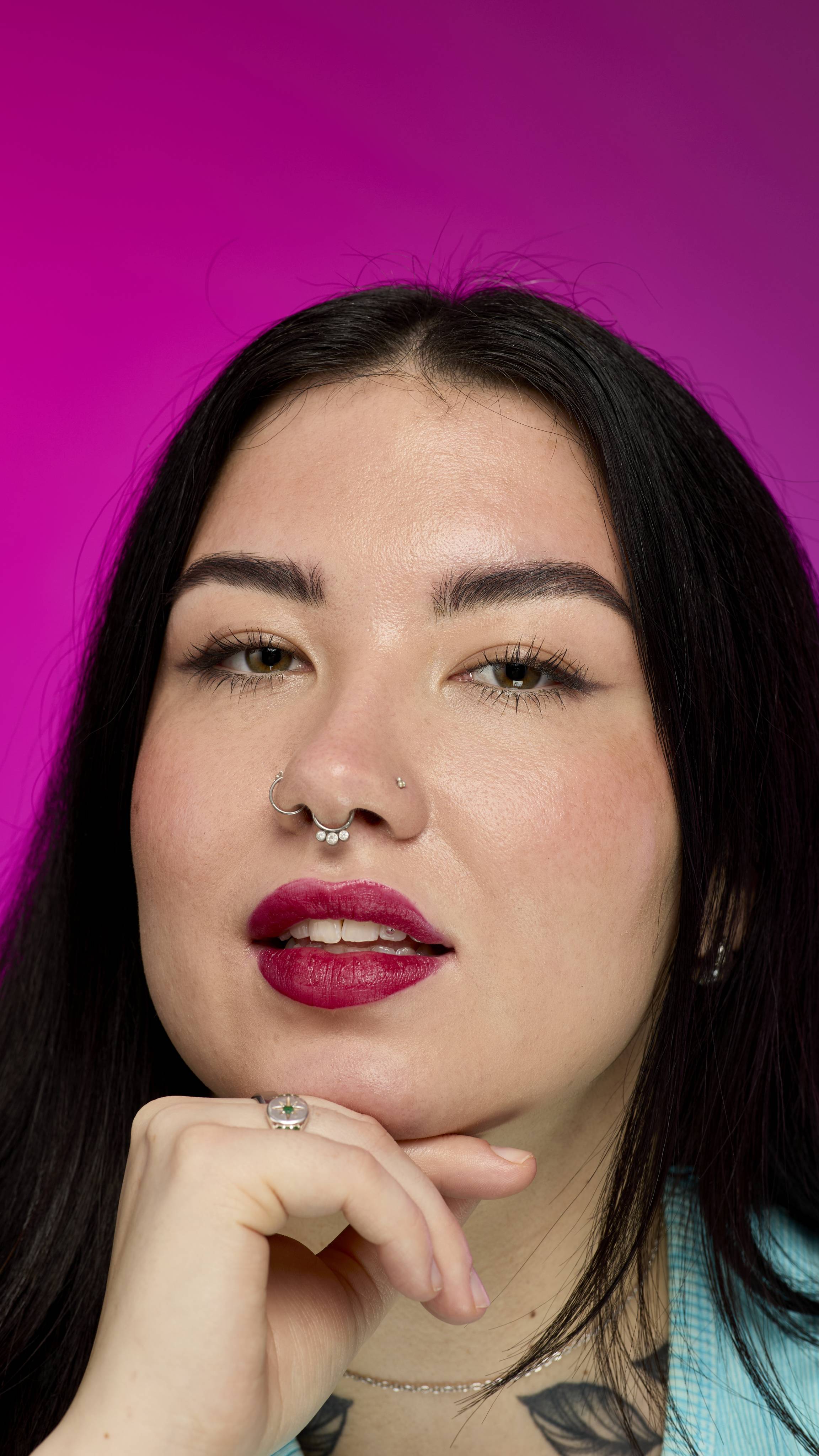 A smiling person with long, dark hair, wearing a yellow top, is shown from the chest up. They are on a vibrant pink background, showing off the deep red Banger lipstick with a subtle shimmer. 