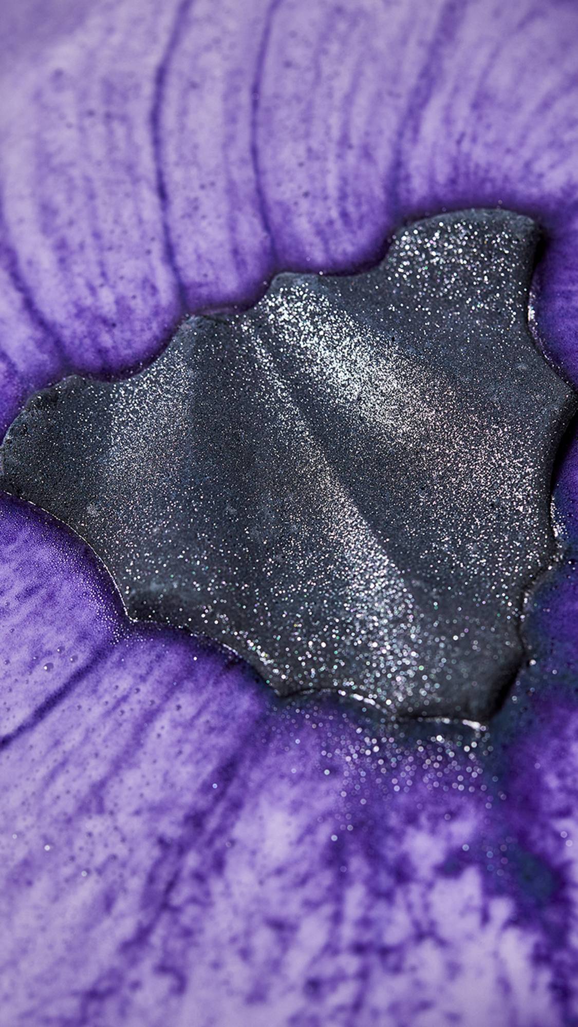 Bat Art. The image shows the Bat Art bath bomb laid on top of the bath water as it gives off a burst of thick, velvety, deep-purple foam as the shimmering coating sparkles under the lighting. 