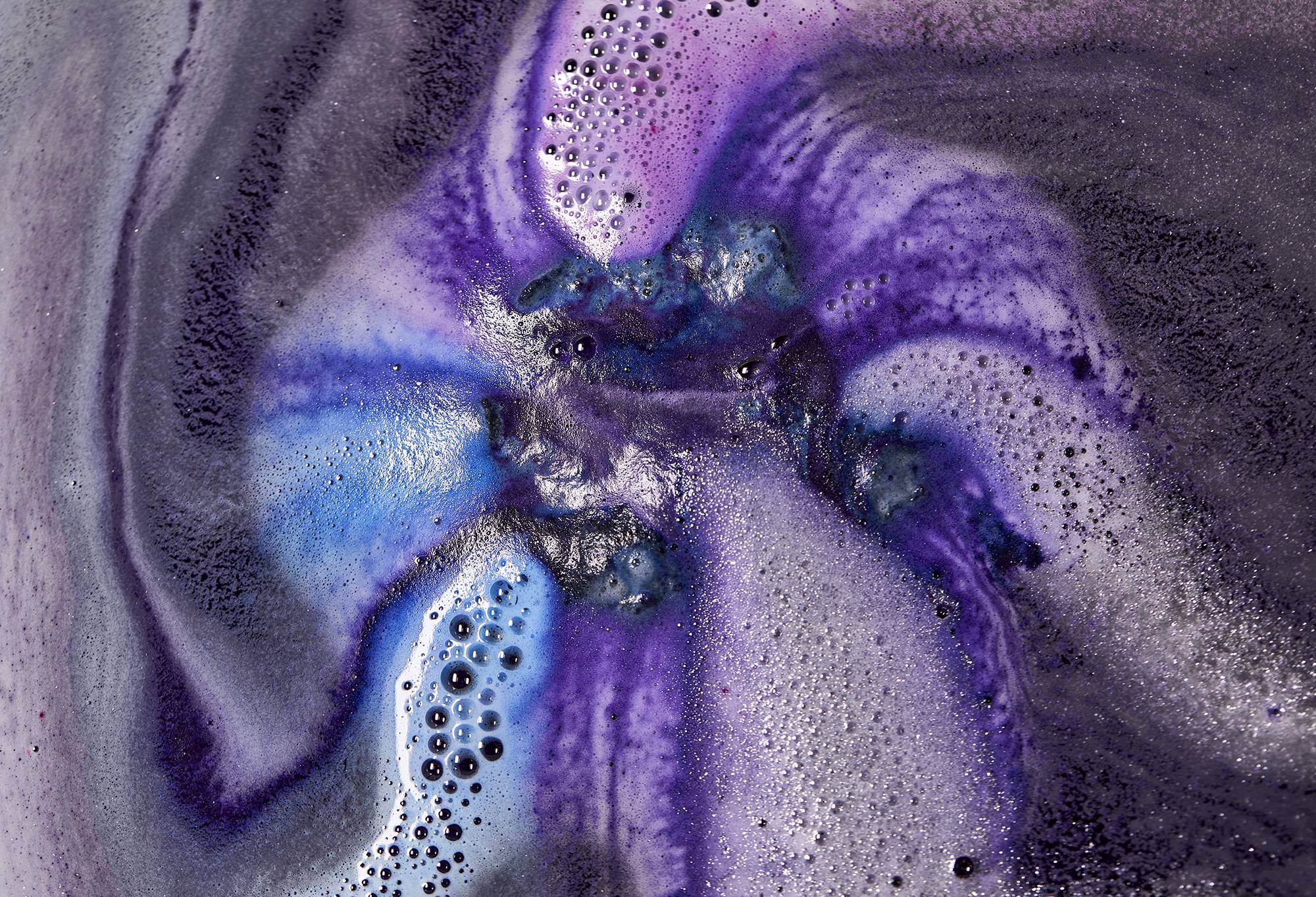 The bath bomb fizzes in the water, releasing dark blue, purple and indigo foam trails across the surface.