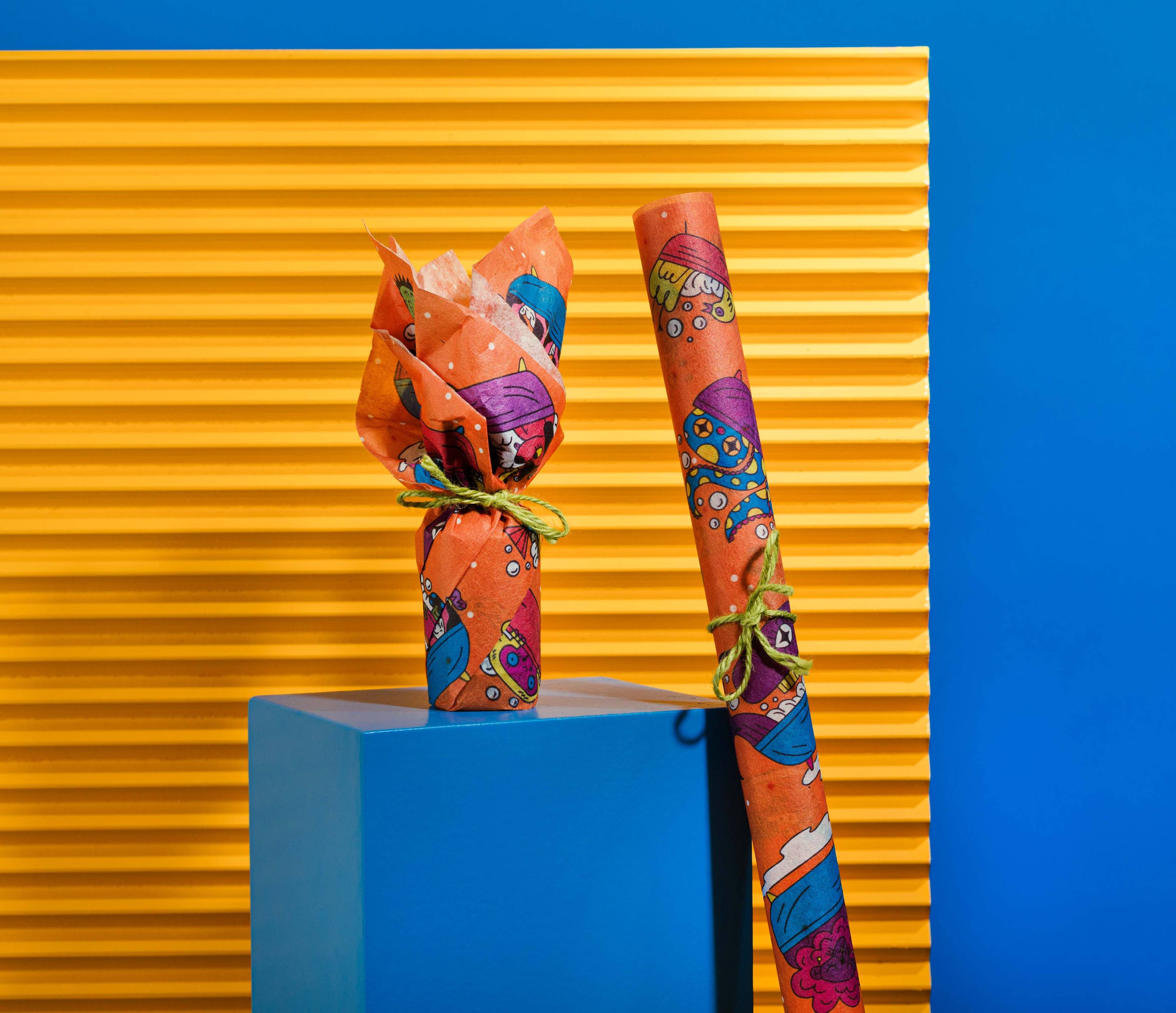 The Lokta paper is wrapped around a product, and rolled up and tied with string, on a blue plinth before an orange background.