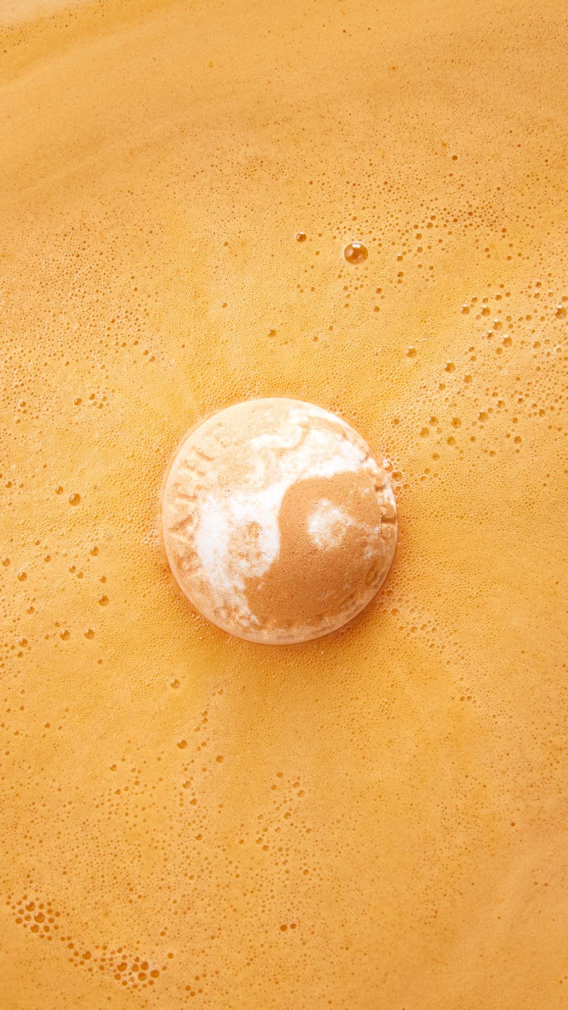 An aerial view of the LUSH "Bather's Digest" orange and white marbled bath bomb dissolving in bright orange bathwater, floating in the centre as it creates a ring of fine foam and bubbles around itself.