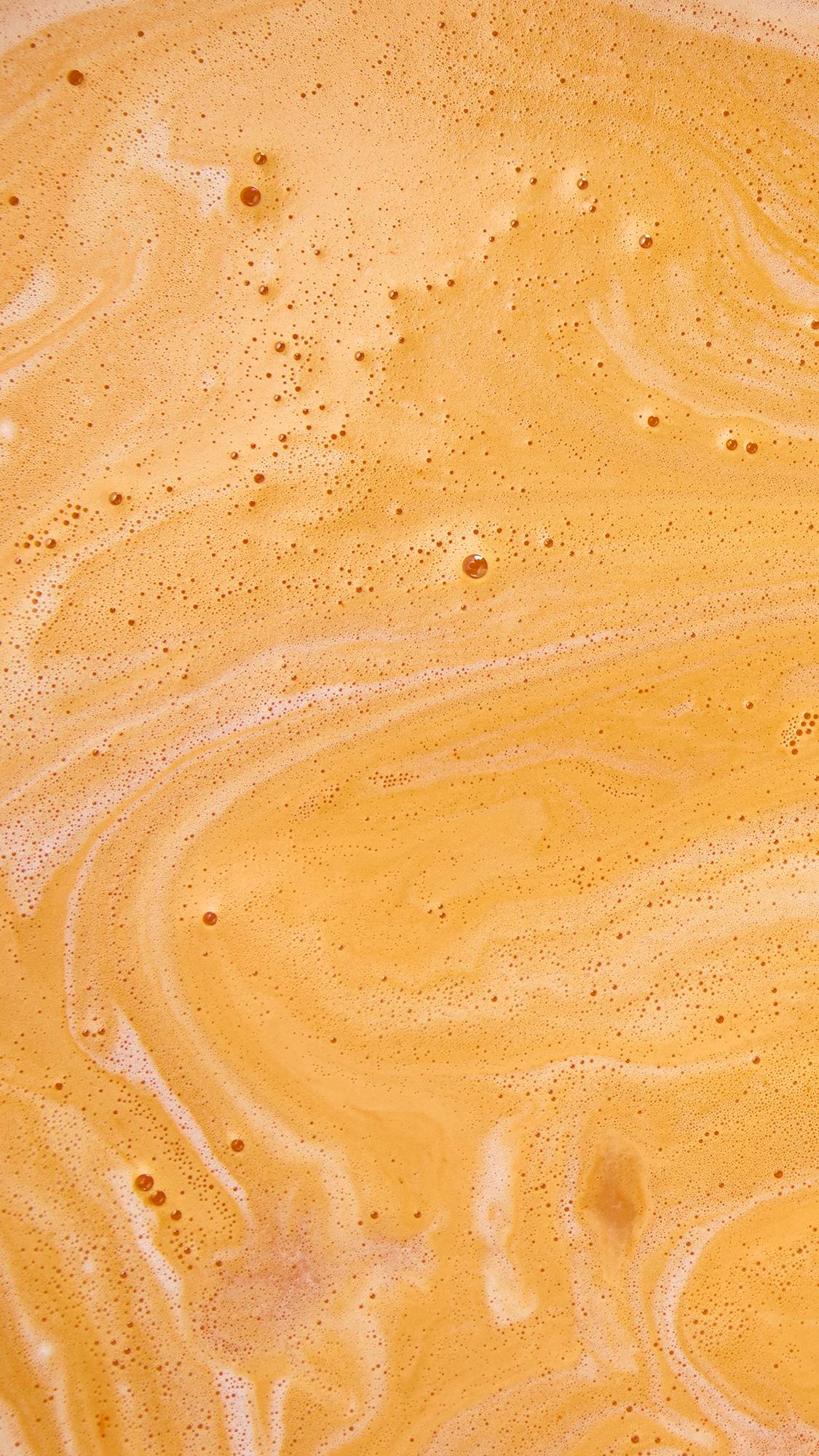 An aerial close-up of vibrant orange bathwater with swirling patterns of creamy white foam and tiny bubbles spread across the surface, made by the LUSH "Bather's Digest" bath bomb.