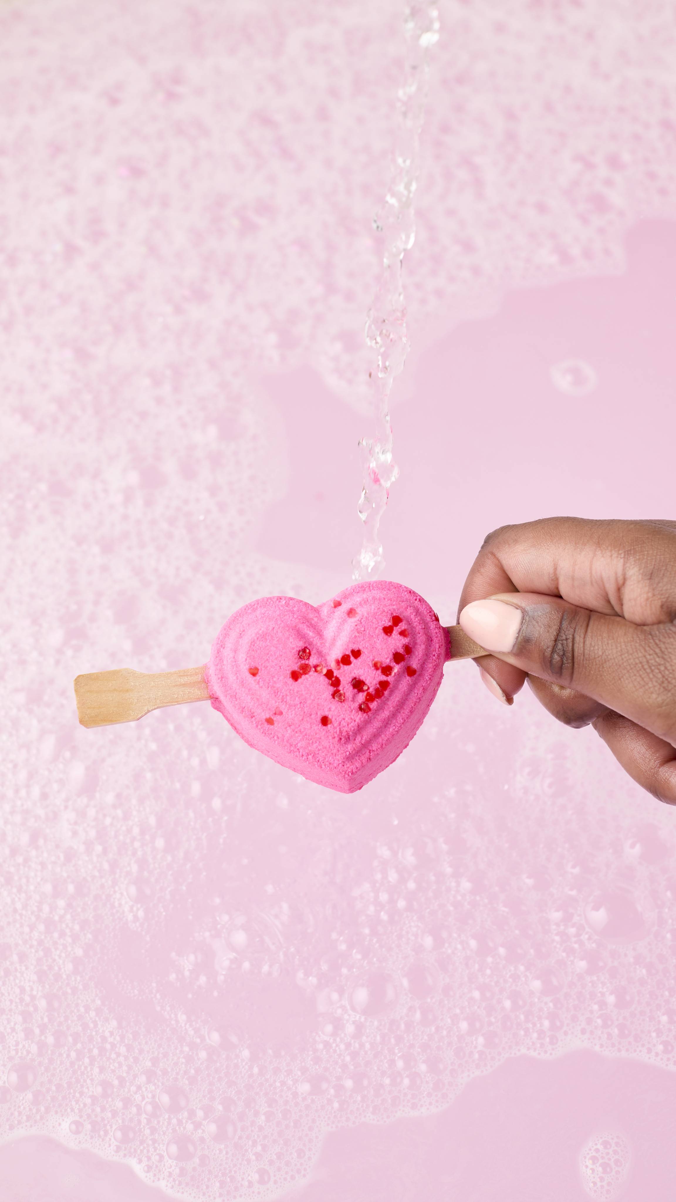 The image shows a hand holding the pink heart-shaped Be My Valentine bubble bar by the wooden stick under a stream of water. The product is held over a tub of pale pink, bubbly bathwater as it begins to dissolve.