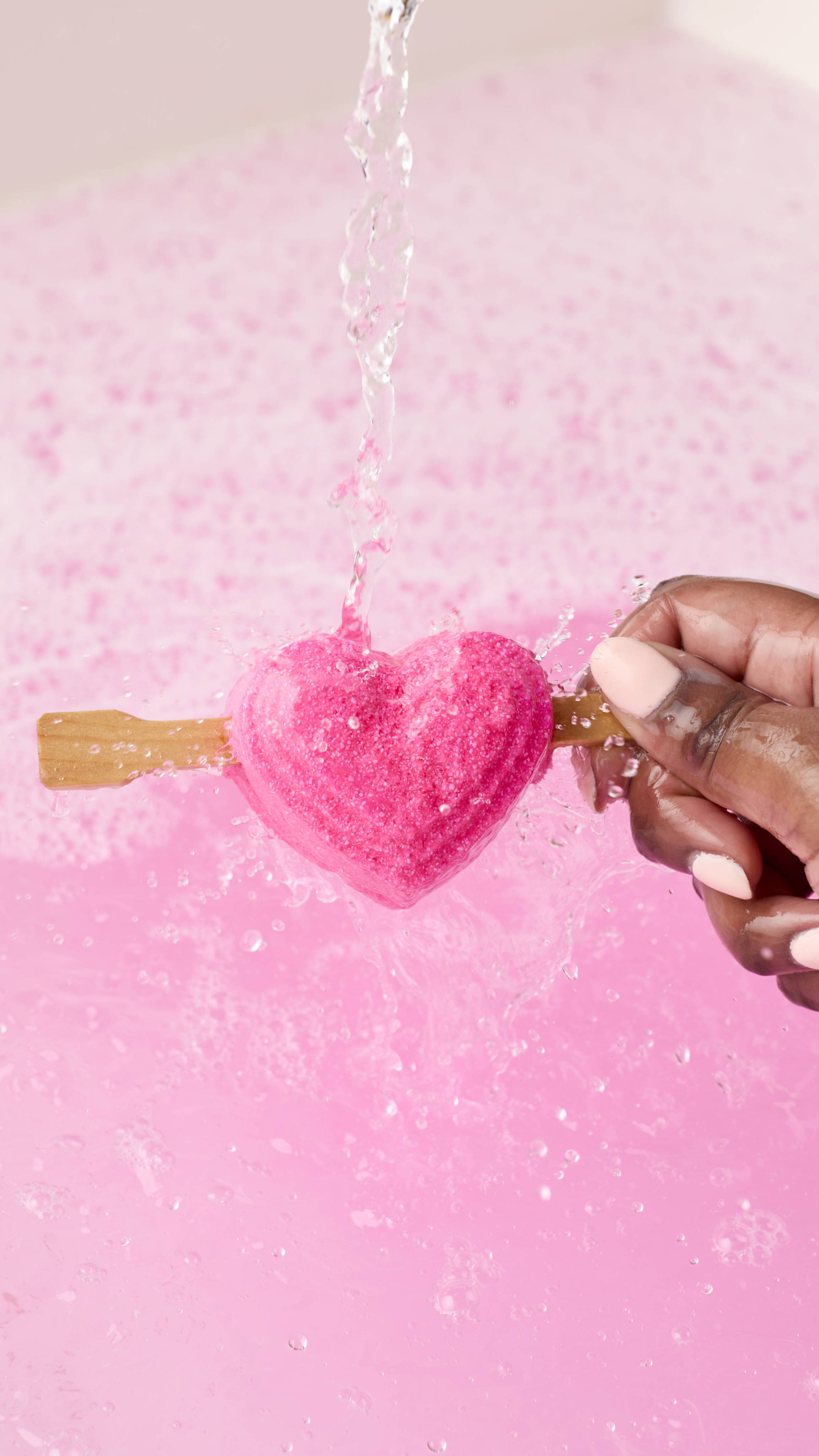 A hand holds the pink heart-shaped Be My Valentine bubble bar on a stick under a stream of water. The bubble bar is held over a tub of pale pink, bubbly bathwater as it begins to dissolve, creating a deeper pink. 