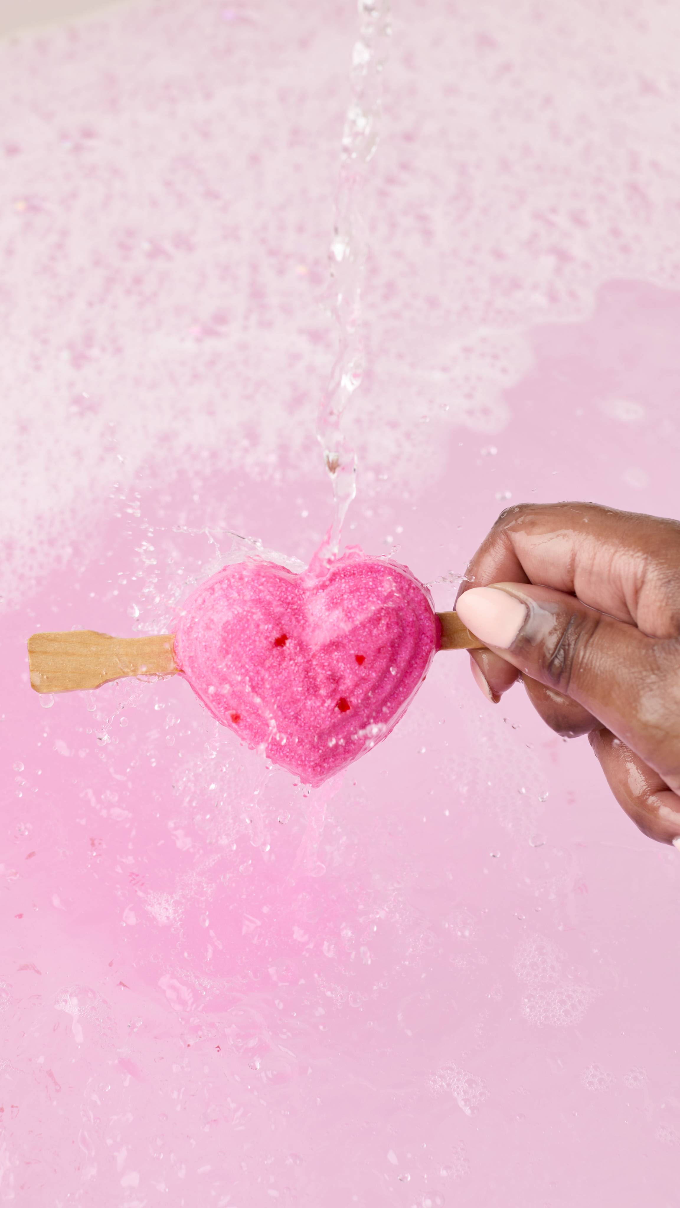 A hand holds the pink heart-shaped Be My Valentine bubble bar on a stick under a stream of water. The bubble bar is held over a tub of pale pink, bubbly bathwater as it begins to dissolve, creating a deeper pink. 