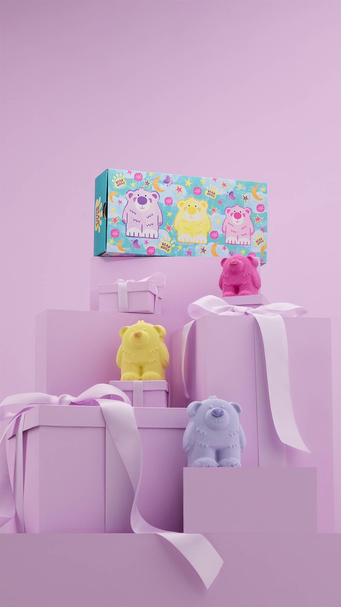 The image shows a large pile of differently-sized wrapped pink gifts with ribbons, on a matching pink background. The colourful Bear Hugs gift box sits on top of the pile, and the three bear-shaped bath bombs sit on other lower pink gifts.