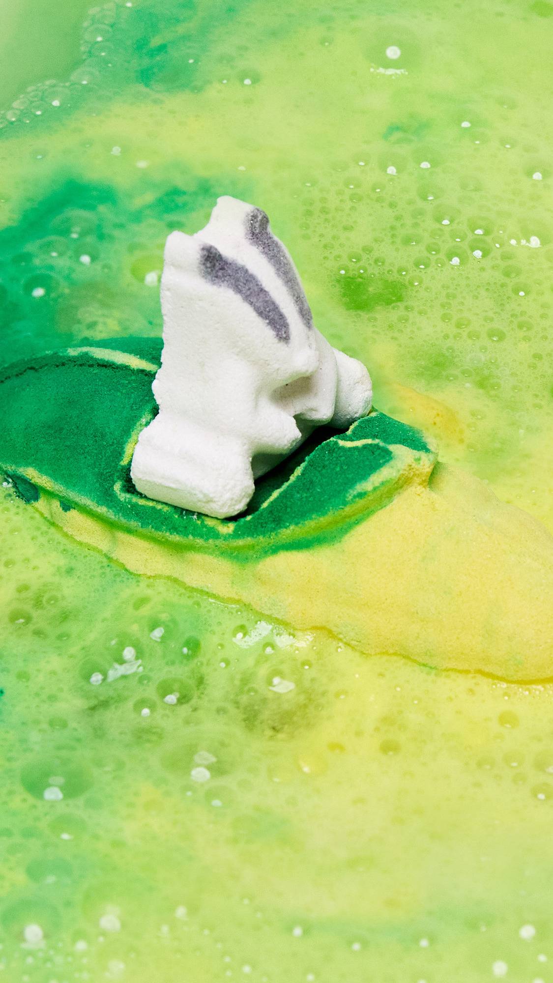 The Beat The Clock Badger bath bomb is sitting perfectly on the bath water as it gives off a blanket of vivid yellow and green swirling foam. 