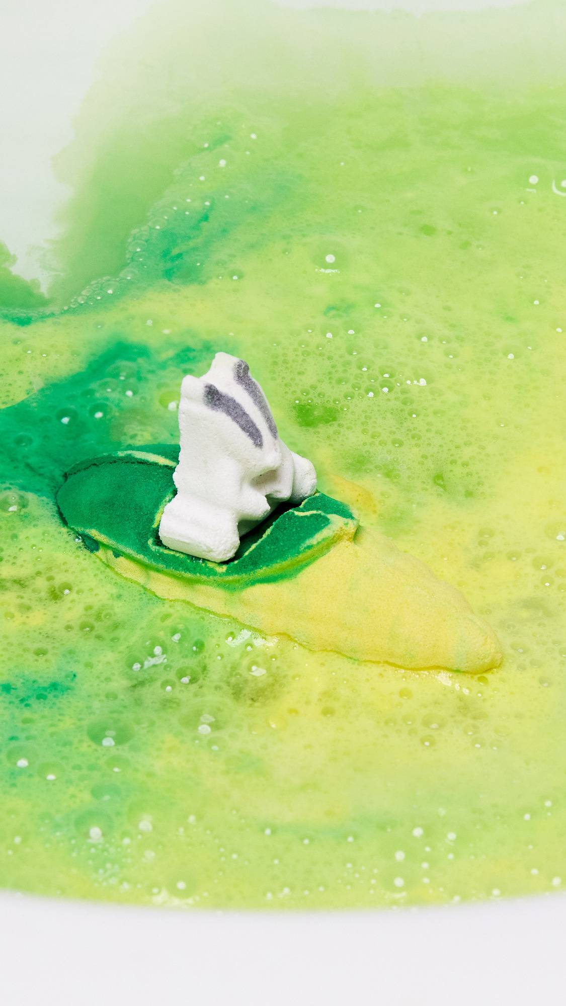 The Beat The Clock Badger bath bomb is sitting perfectly on the bath water as it gives off a blanket of vivid yellow and green swirling foam. 