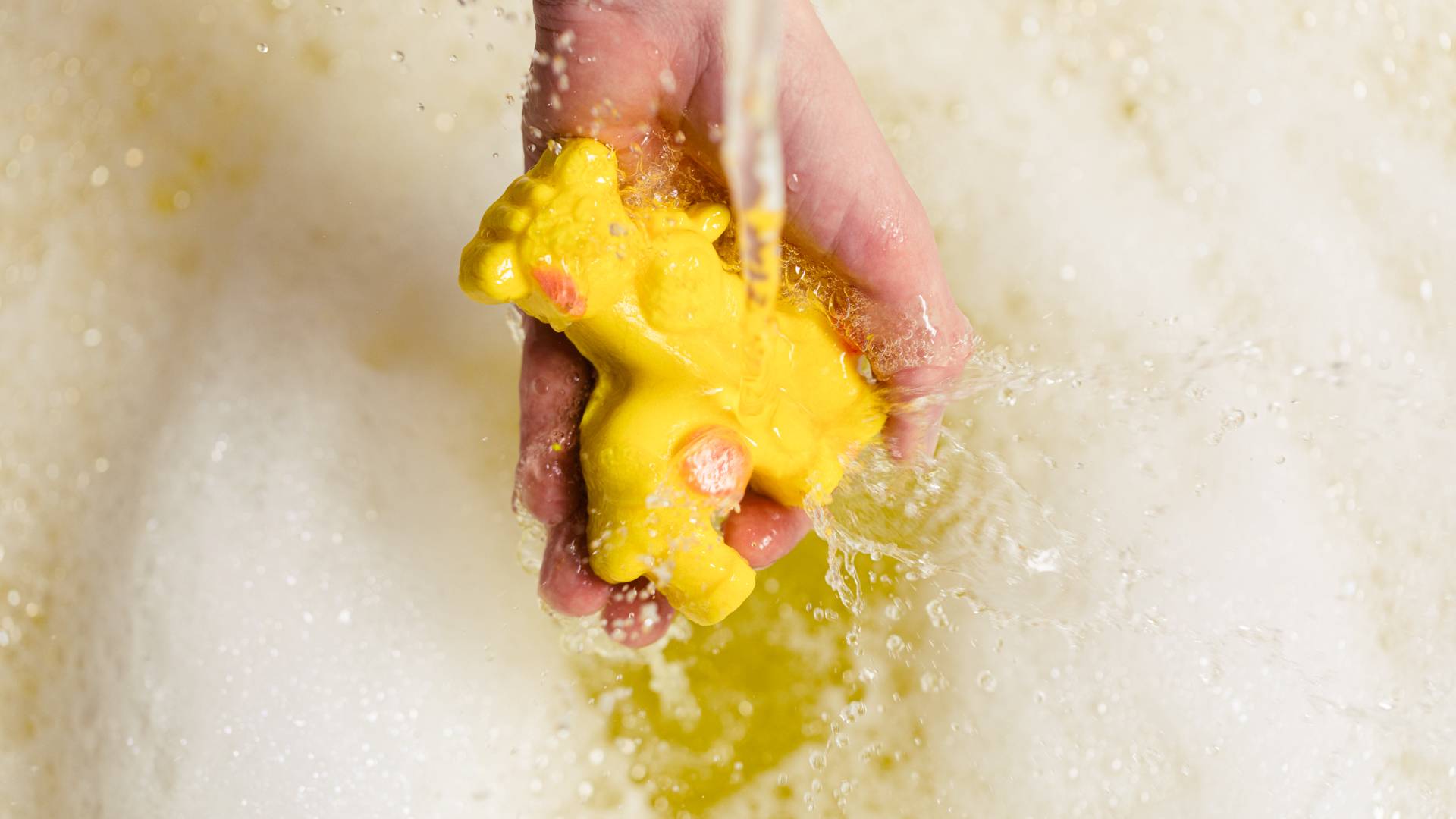 Yellow giraffes with pink spots being held under a running tap, to create bubbles in yellow water below.