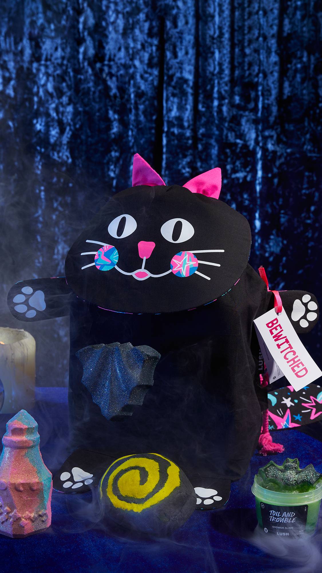 The image shows the Bewitched, cat-shaped gift bag on a dark blue, velvet background. The four Halloween LUSH products are beside it. 