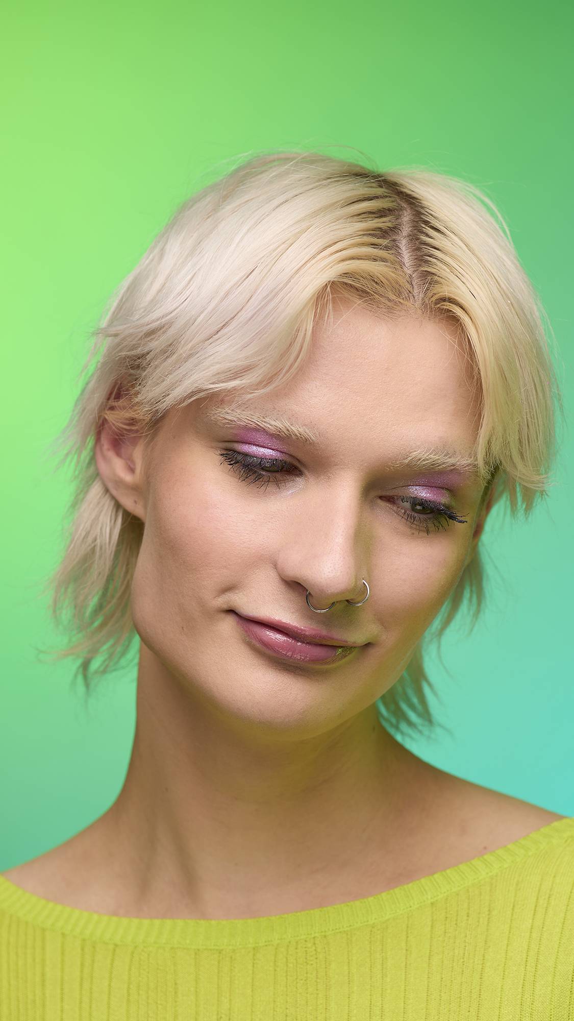 The image shows a close-up of the model's face on a pale blue-to-green gradient background. The model's make-up is done with the focal point being a subtle, rose-pink, shimmering eyeshadow made by the Big Bang Glitter Crumble. 