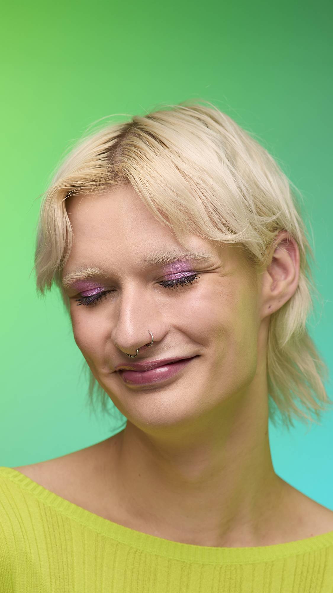 The image shows a close-up of the model's face on a pale blue-to-green gradient background. The model's make-up is done with the focal point being a subtle, rose-pink, shimmering eyeshadow made by the Big Bang Glitter Crumble. 